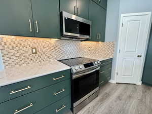 Kitchen featuring green cabinetry, appliances with stainless steel finishes, light stone countertops, tasteful backsplash, and light wood finished floors