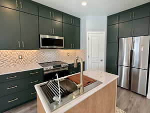 Kitchen with stainless steel appliances, light wood-type flooring, backsplash, green cabinets, and light stone counters