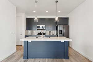 Kitchen with backsplash, a kitchen island with sink, stainless steel appliances, hanging light fixtures, and light wood-style floors