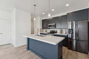 Kitchen featuring appliances with stainless steel finishes, a kitchen island with sink, decorative backsplash, decorative light fixtures, and light wood-style floors