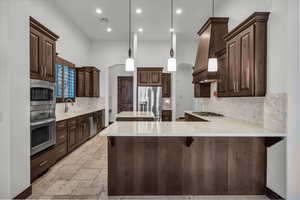 Kitchen featuring dark brown cabinetry, backsplash, appliances with stainless steel finishes, a peninsula, and light stone countertops