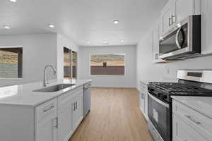 Kitchen featuring appliances with stainless steel finishes, white cabinets, light stone countertops, light wood finished floors, and a center island with sink