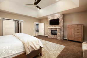 Carpeted bedroom featuring a barn door, a ceiling fan, a stone fireplace, and a tray ceiling