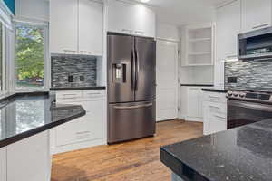 Kitchen with appliances with stainless steel finishes, open shelves, decorative backsplash, light wood finished floors, and dark stone countertops