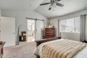 Bedroom with vaulted ceiling, a barn door, light colored carpet, ensuite bath, and a ceiling fan