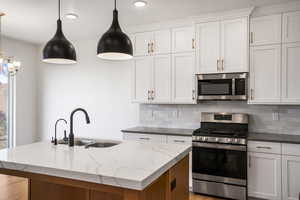 Kitchen featuring appliances with stainless steel finishes, decorative light fixtures, backsplash, a center island with sink, and white cabinetry