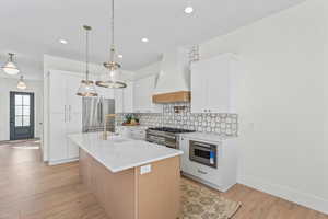 Kitchen with a kitchen island with sink, premium range hood, light wood-style floors, white cabinets, and decorative backsplash