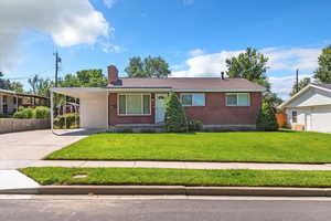 Single story home featuring concrete driveway, brick siding, a front yard, a chimney, and an attached carport