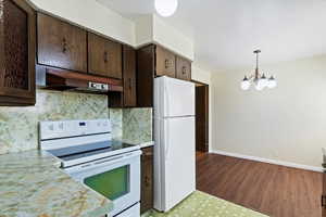 Kitchen with white appliances, light countertops, dark brown cabinets, under cabinet range hood, and hanging light fixtures