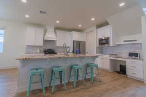 Kitchen with white cabinets, a kitchen breakfast bar, appliances with stainless steel finishes, custom exhaust hood, and light stone counters