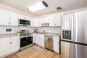 Kitchen with appliances with stainless steel finishes, white cabinetry, light wood finished floors, tasteful backsplash, and light stone countertops