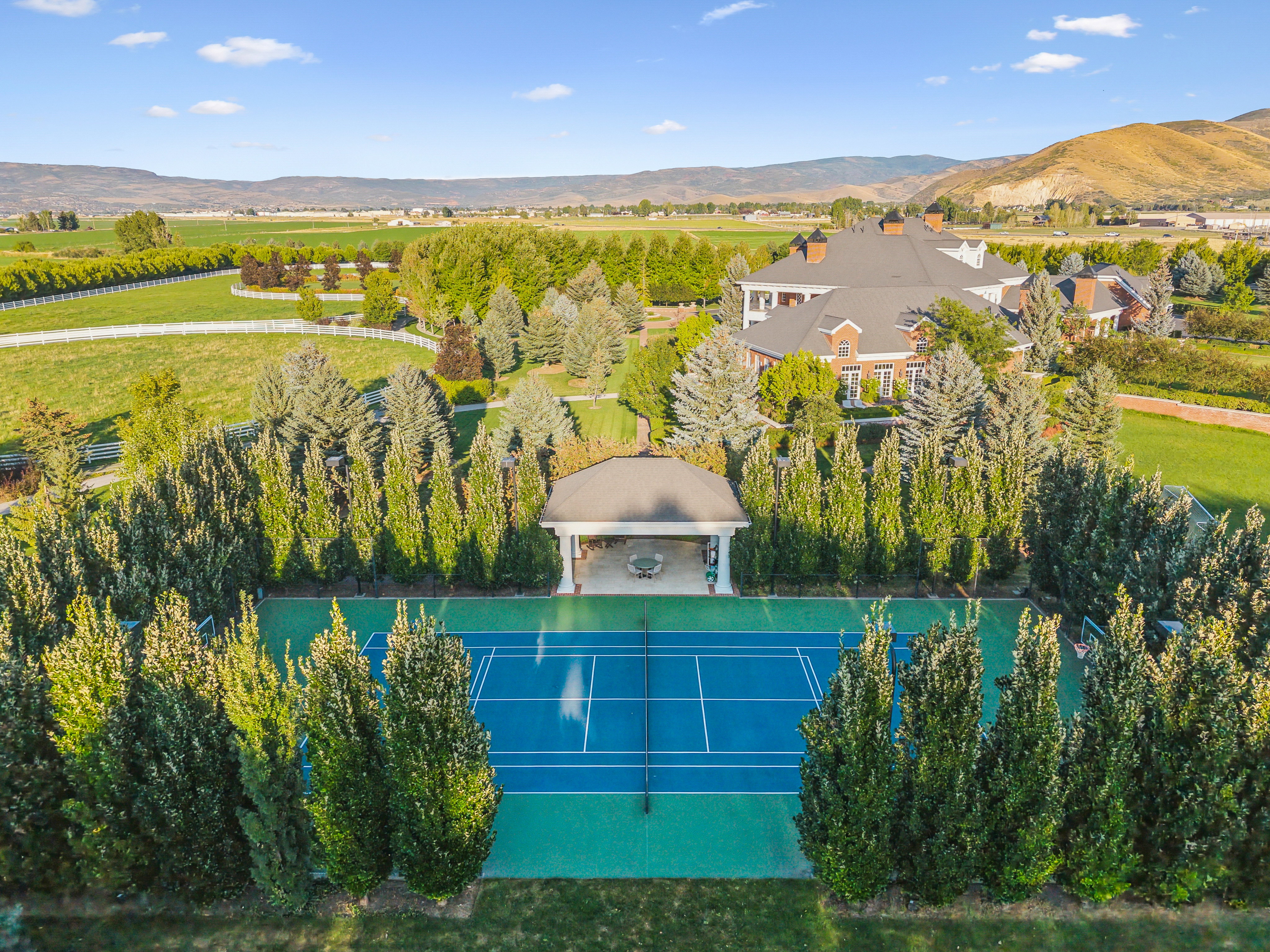 Full-size tennis court with a blue playing surface and green surrounds, bordered by mature evergreen trees