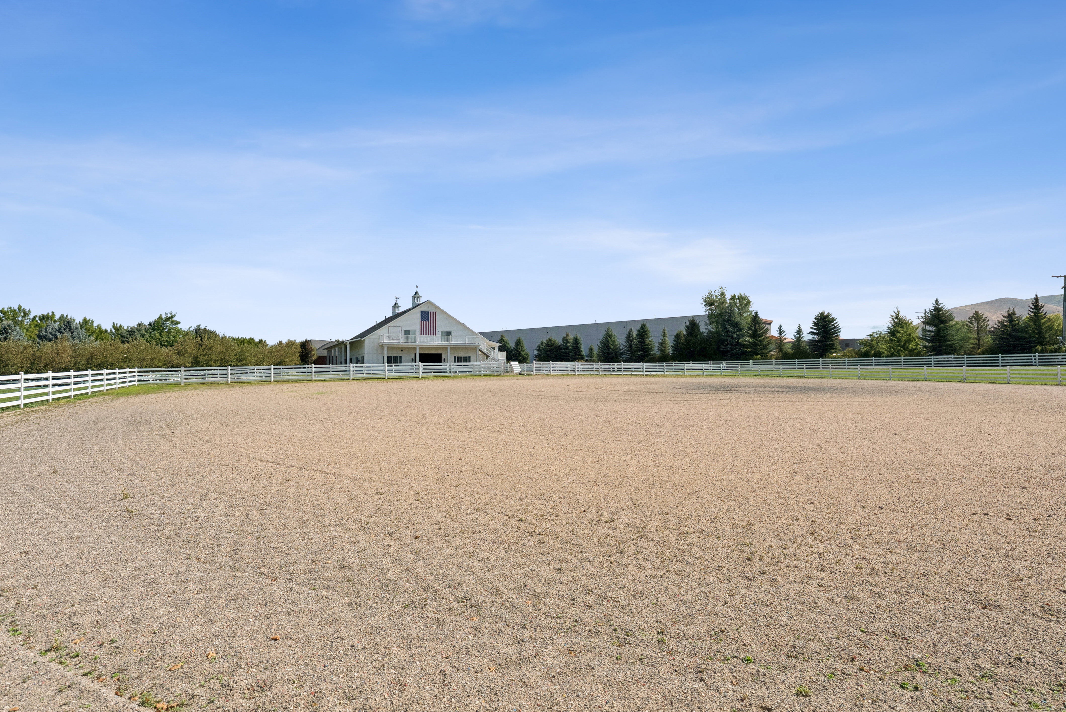 Expansive outdoor riding arena with a white perimeter fence, featuring a large white barn structure in the background