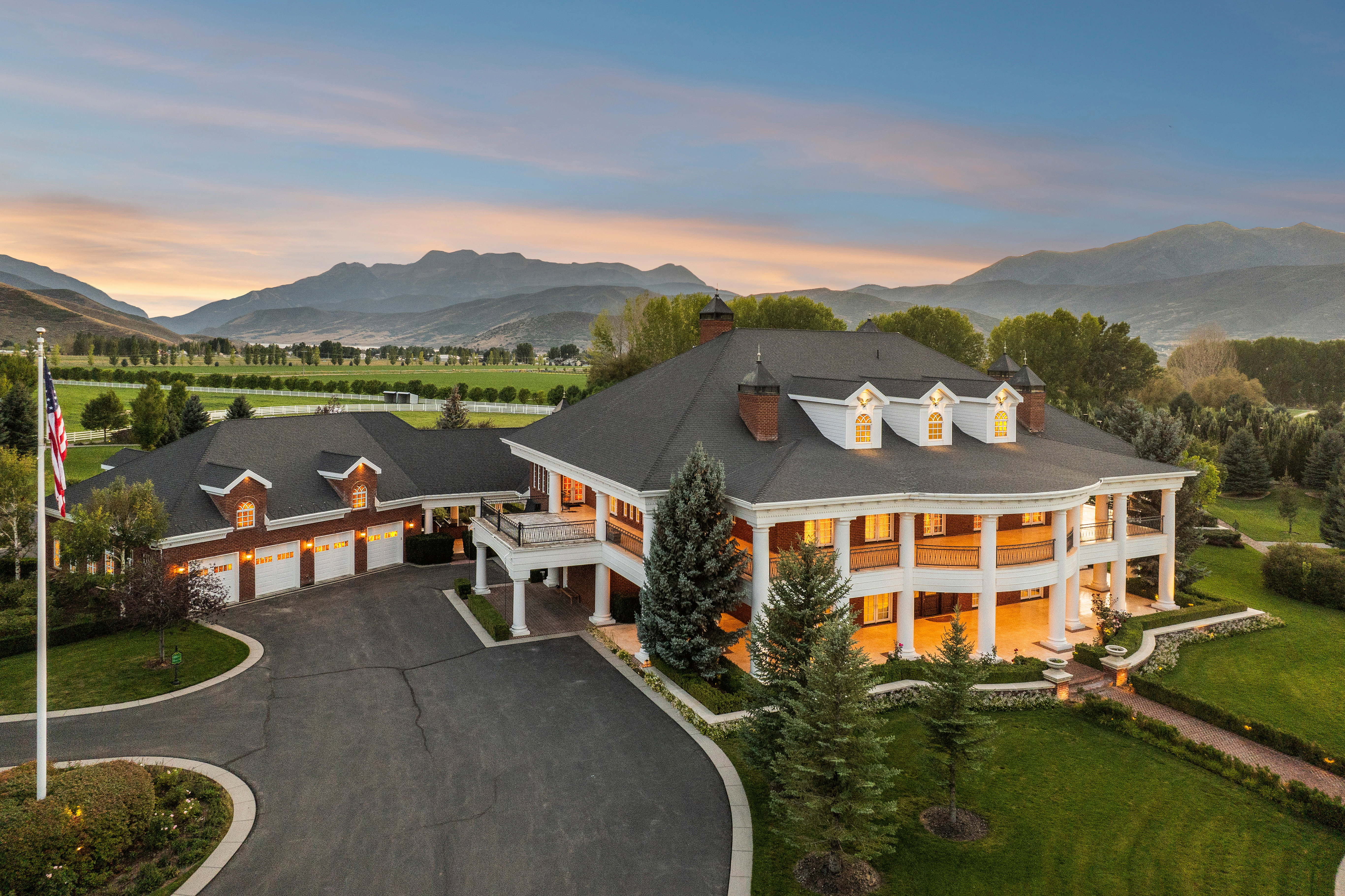 Circular driveway with a grand Colonial-style residence featuring a wrap-around porch supported by white columns, multiple dormer windows, and a multi-car garage with brick accents