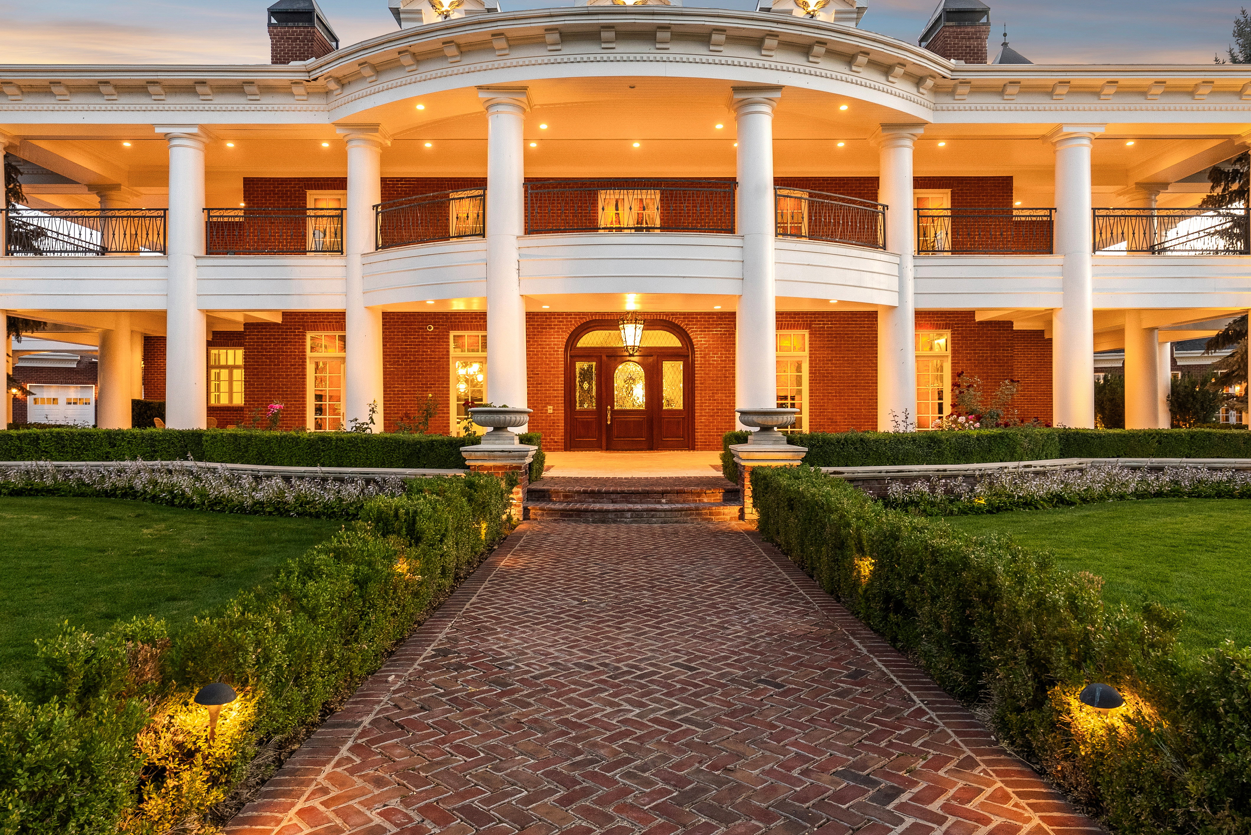 Stately brick residence featuring a grand entrance with a columned portico, a curved second-story balcony with metal railings, and a meticulously landscaped front yard