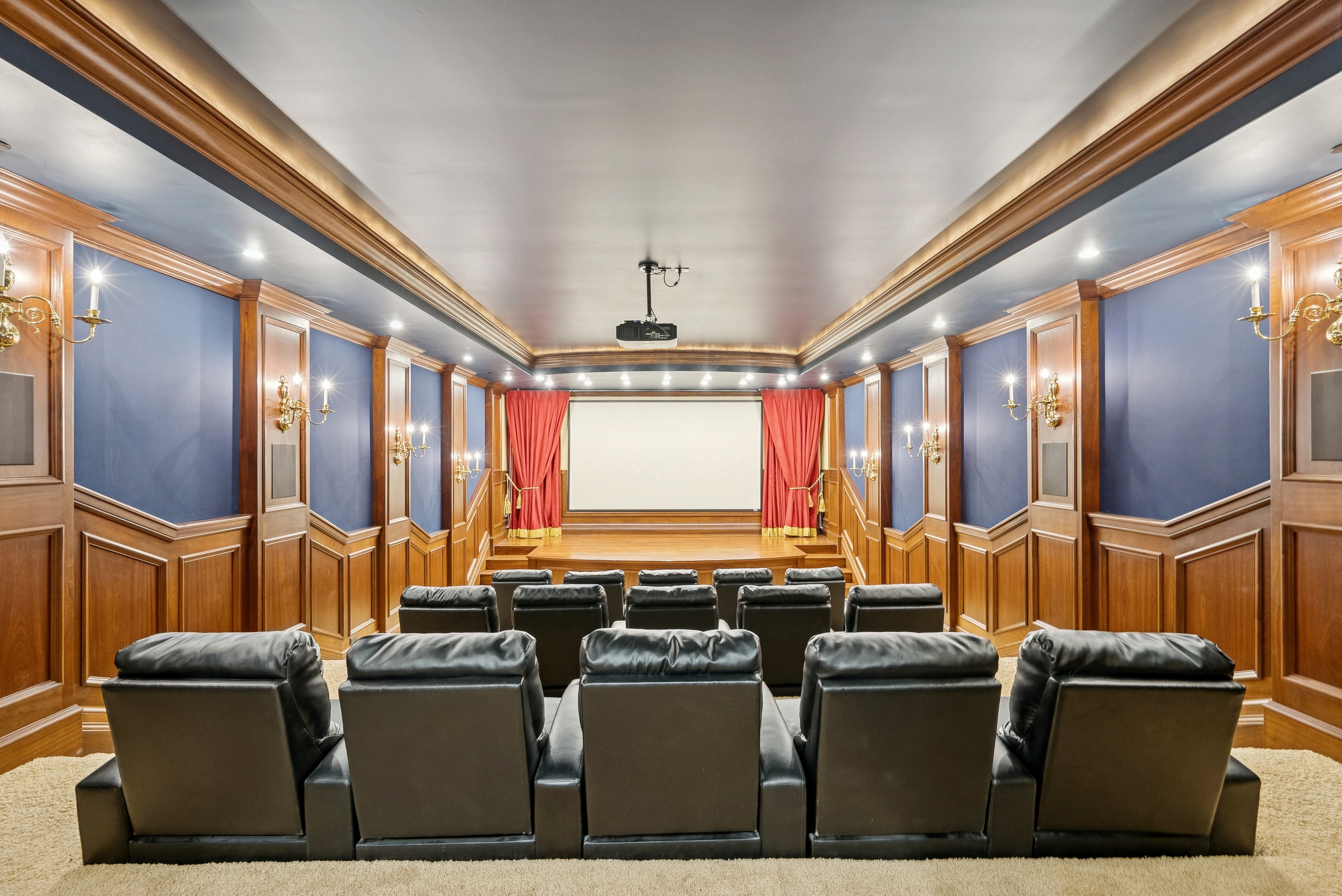 Dedicated home theater featuring tiered seating, wood paneling, sconce lighting, a projection screen, and red stage drapes