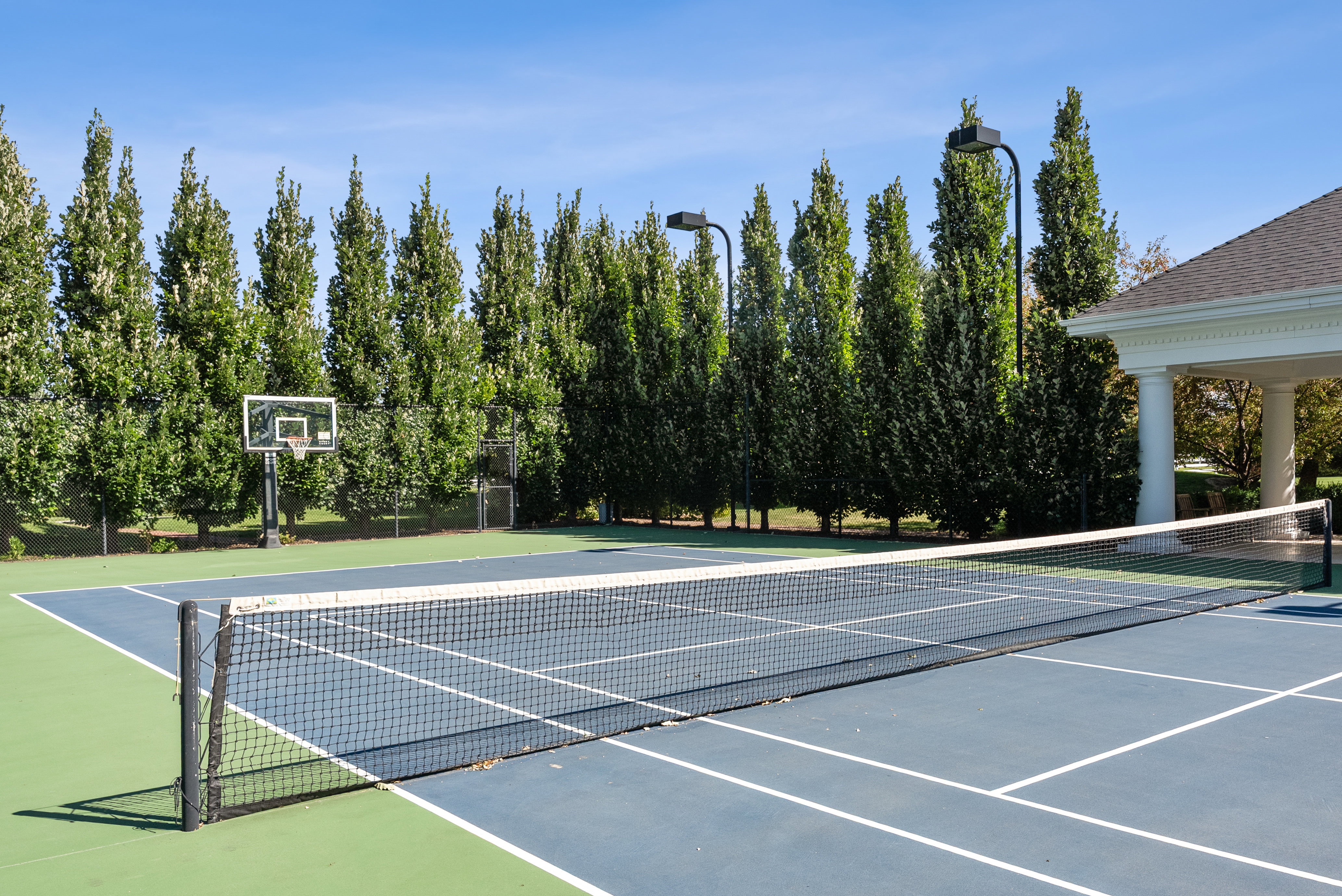 Outdoor multi-sport court featuring a tennis net, basketball hoop, and perimeter lighting