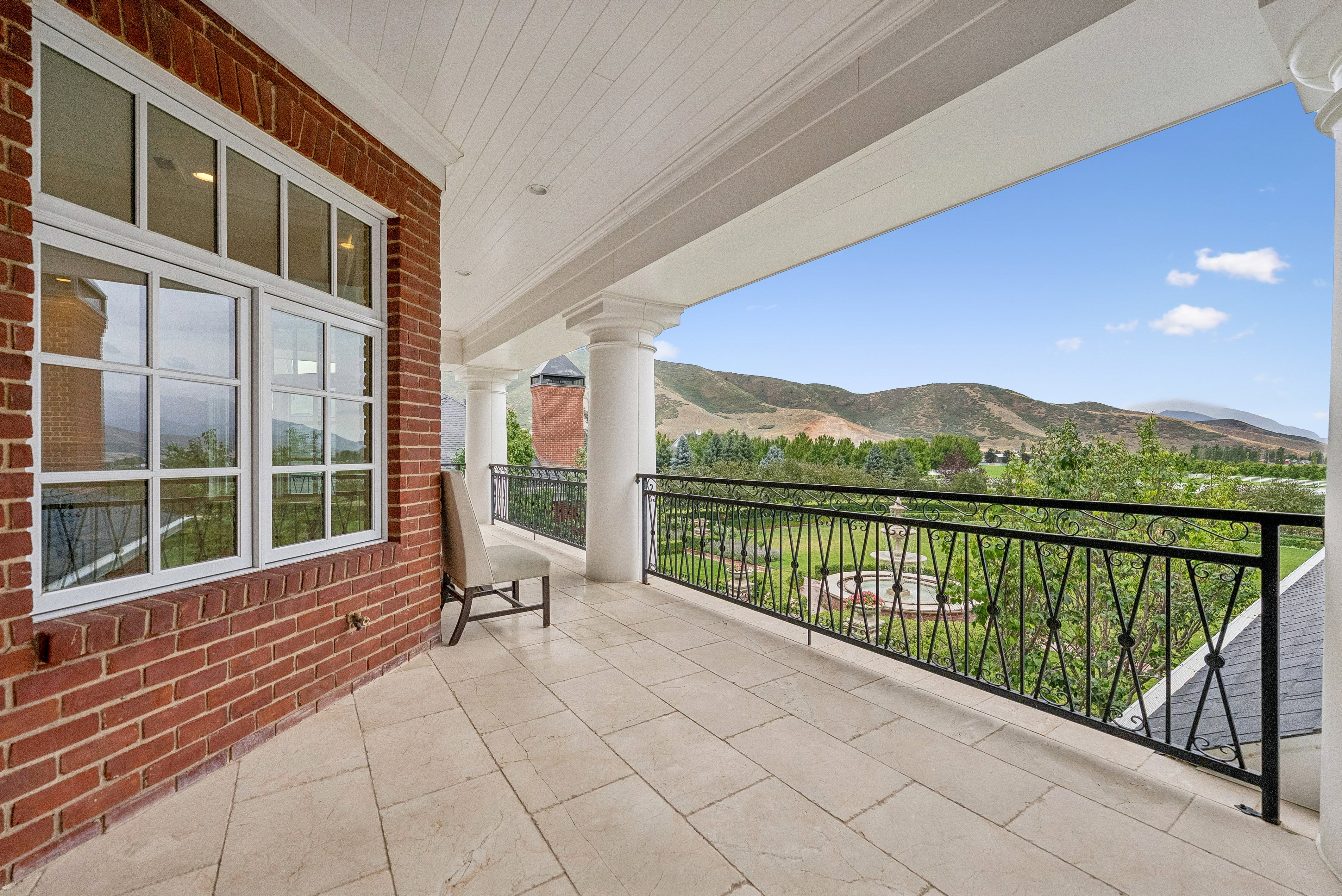 Expansive balcony featuring durable tile flooring, classic white columns, and a decorative wrought-iron railing overlooking a lush landscape with distant mountains