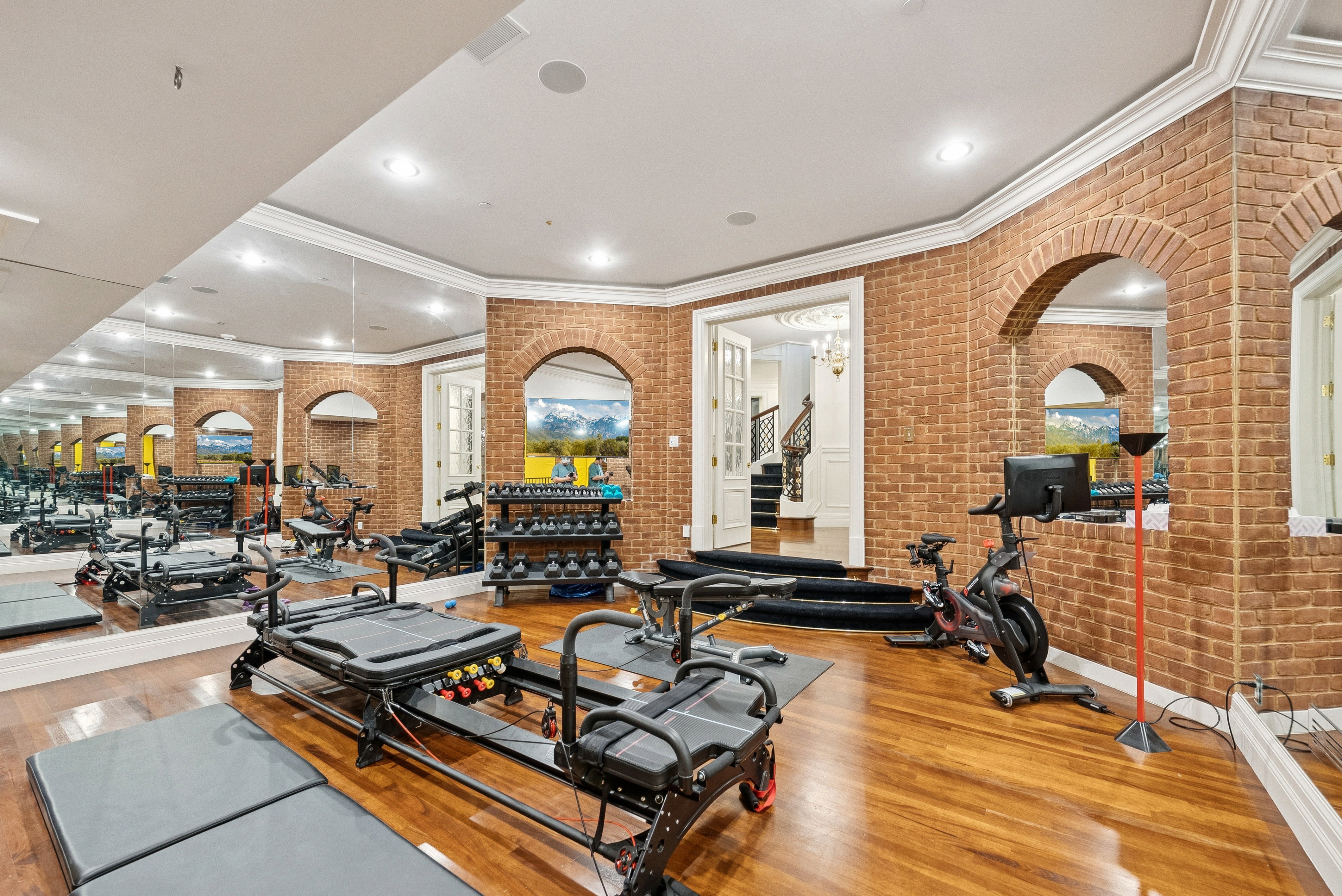 Dedicated fitness room featuring wood-finish flooring, full-wall mirrors, brick-finish walls with arched openings, and crown molding