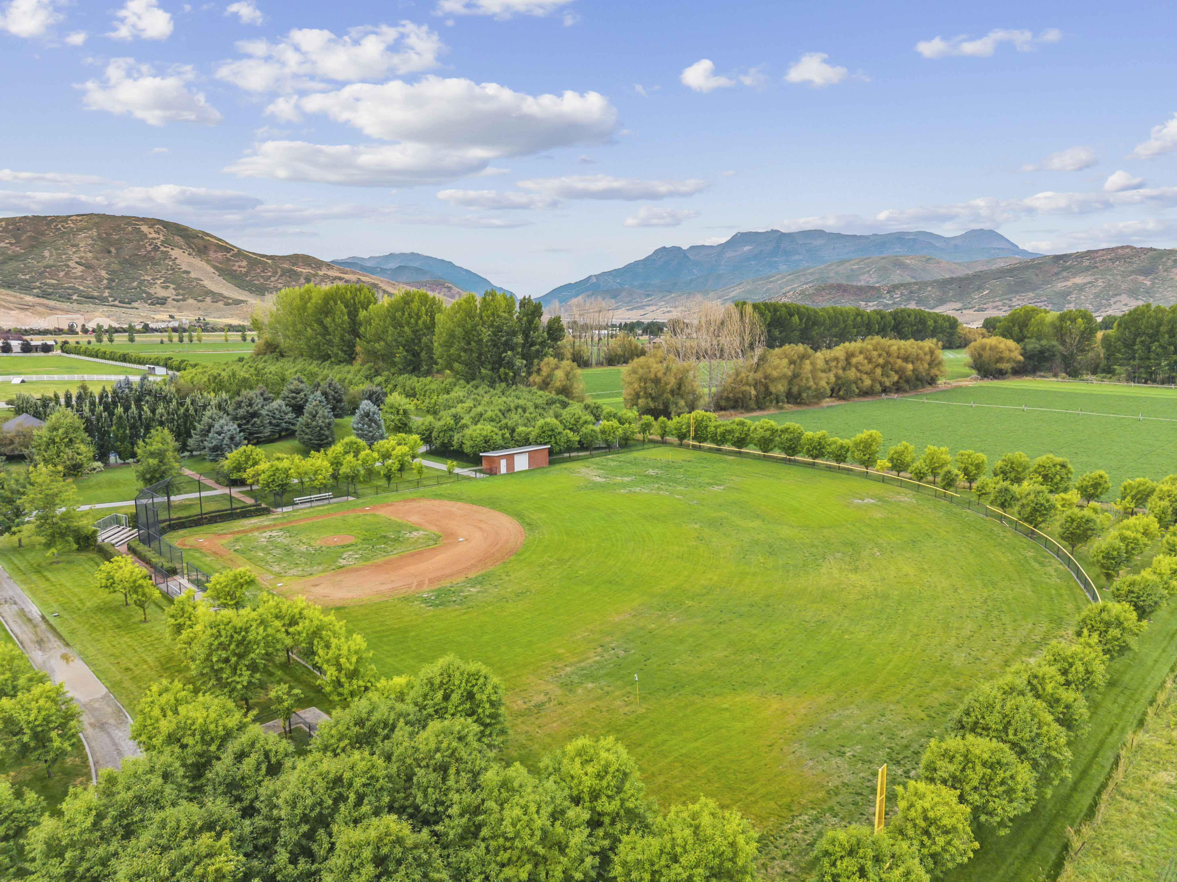 Expansive green field featuring a baseball diamond, surrounded by mature trees, a red and white storage shed, and a mountain backdrop