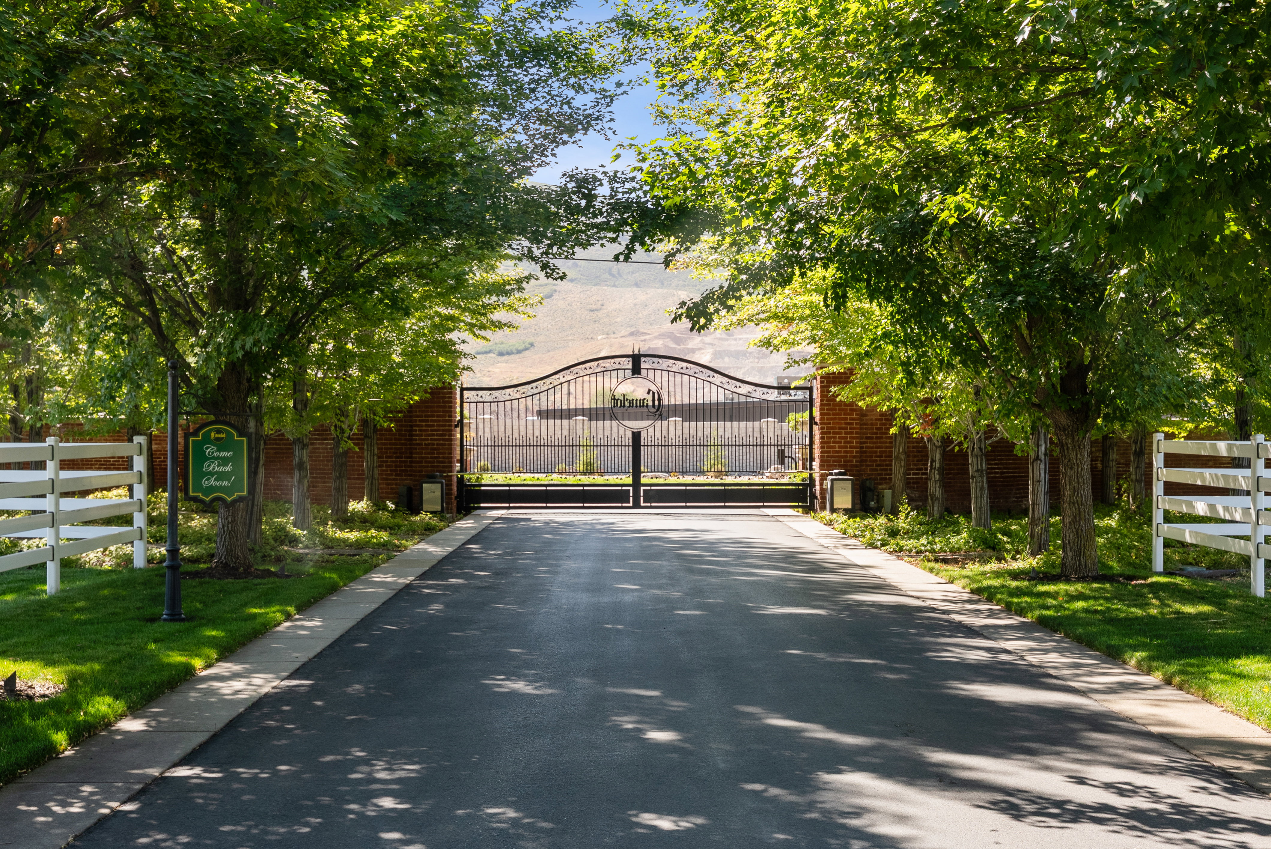 Paved driveway entry featuring an ornate wrought iron gate, brick privacy wall, white split-rail fencing, mature trees, and manicured lawns