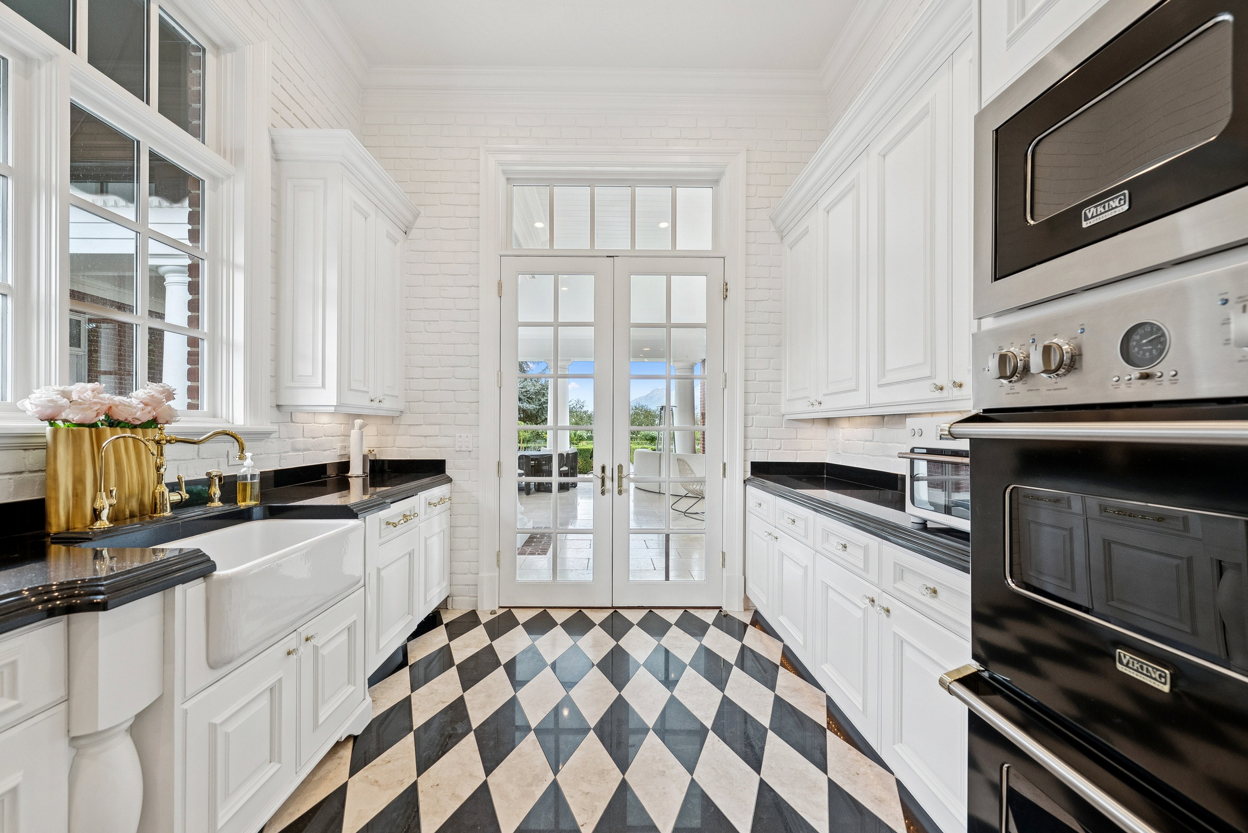 Kitchen featuring black and white diamond-patterned flooring, white shaker-style cabinetry, black countertops, and a farmhouse sink with a brass-finish faucet