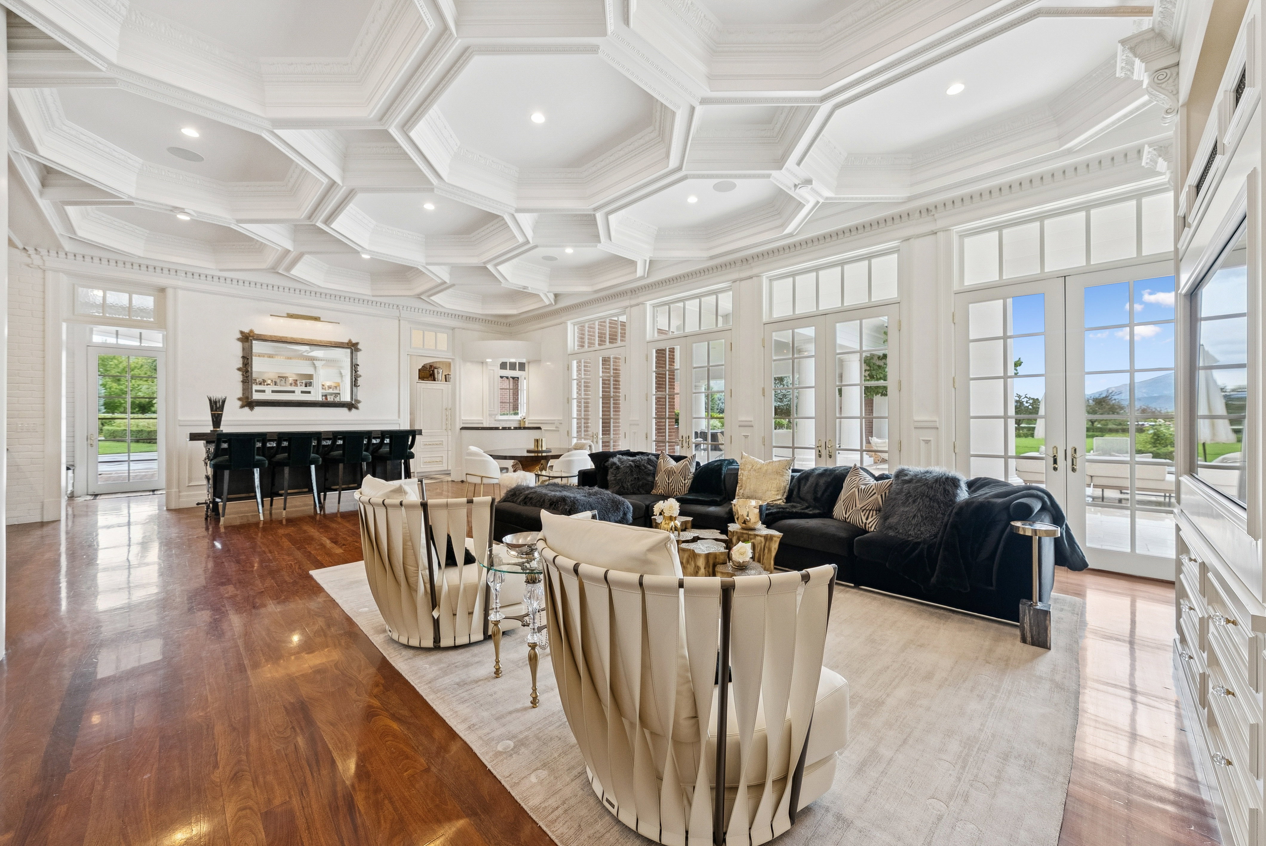 Spacious room featuring a multi-level coffered ceiling with recessed lighting, hardwood flooring, multiple French doors, a built-in wet bar, and extensive white millwork