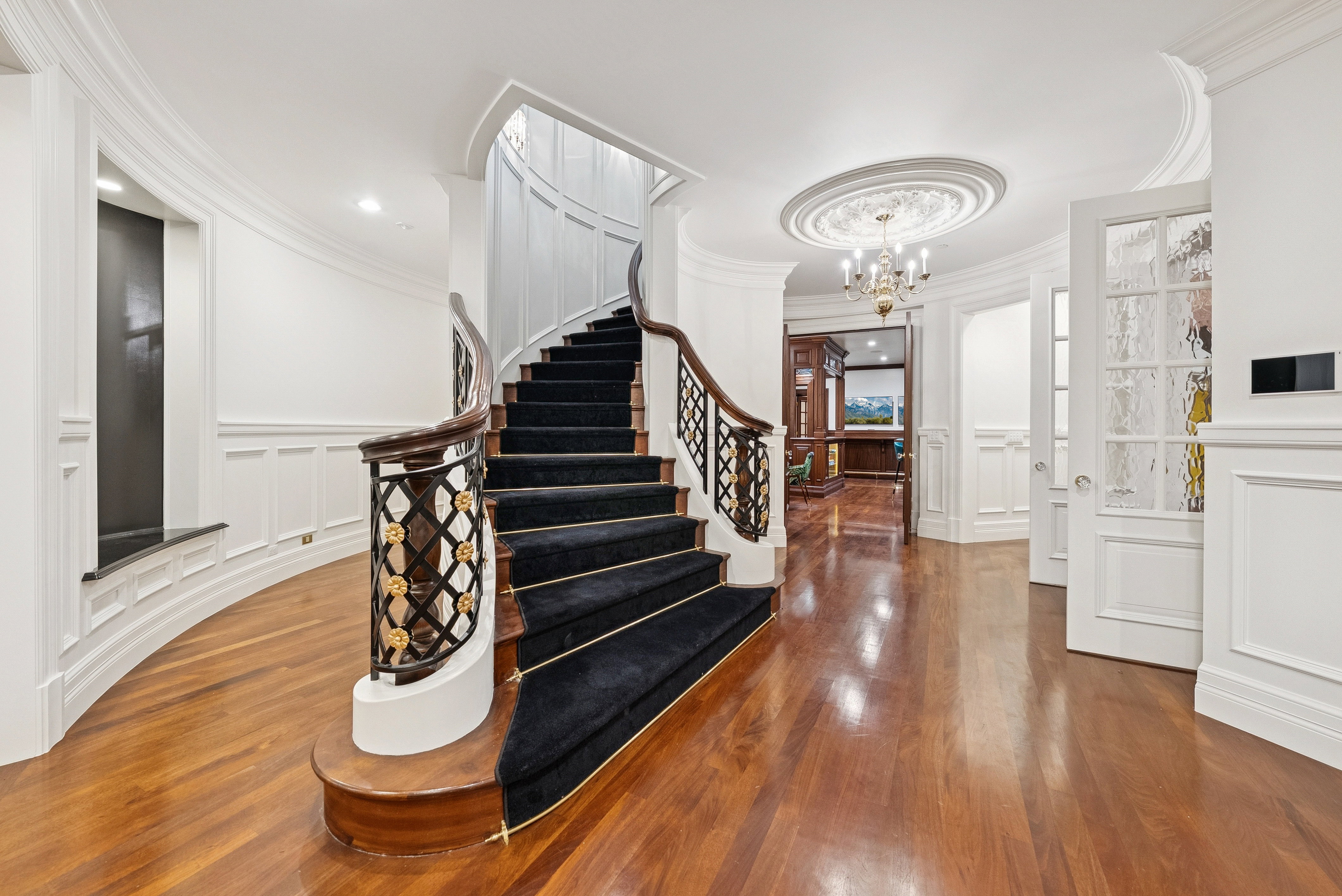 Grand foyer featuring a curved staircase with a dark wood banister and ornate metal balusters, rich wood flooring, wainscoting, decorative ceiling medallion with a chandelier, and a paneled door with textured glass