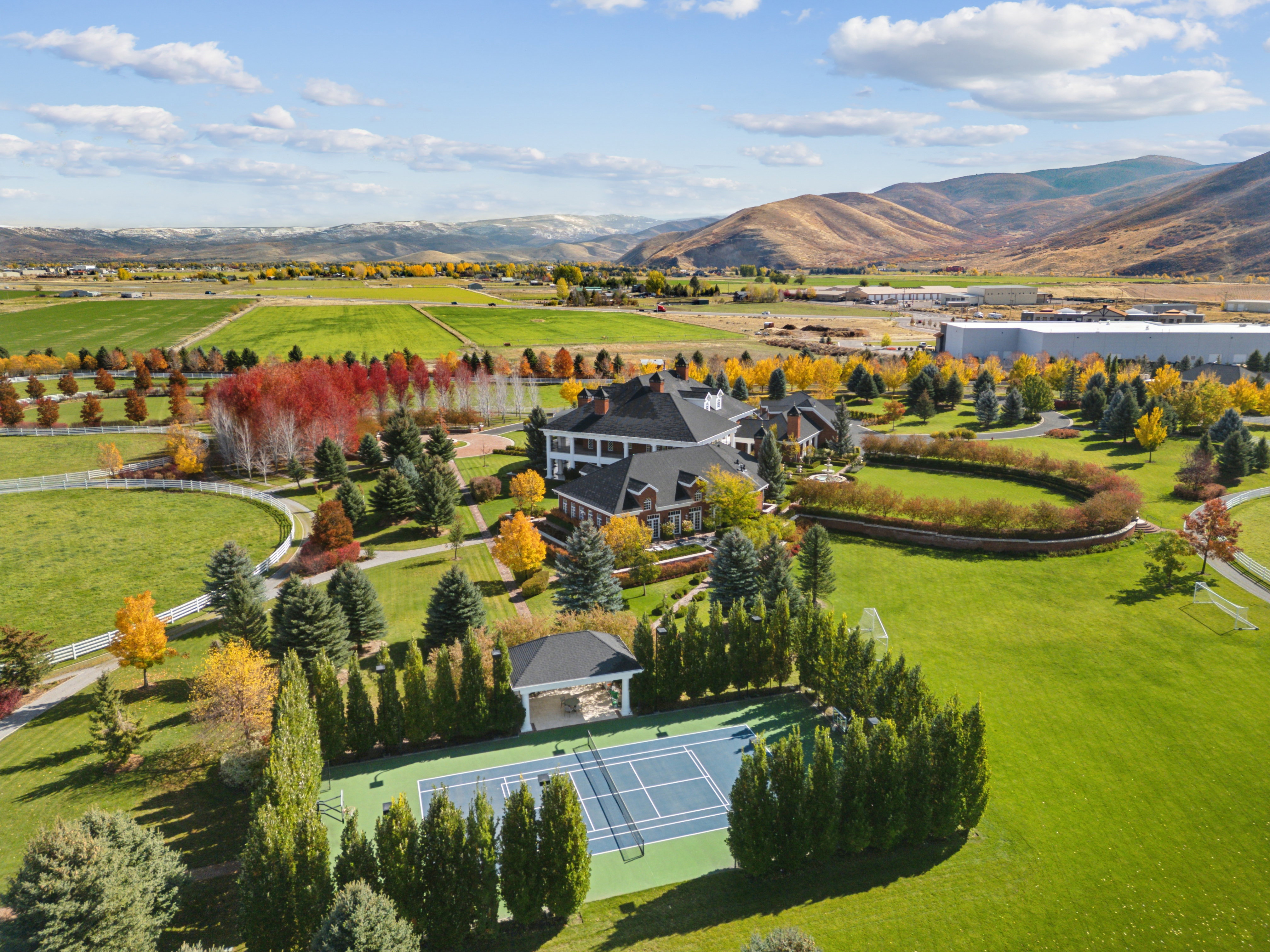 Expansive estate featuring a large main residence, a tennis court with a shaded pavilion, and a surrounding landscape of mature trees
