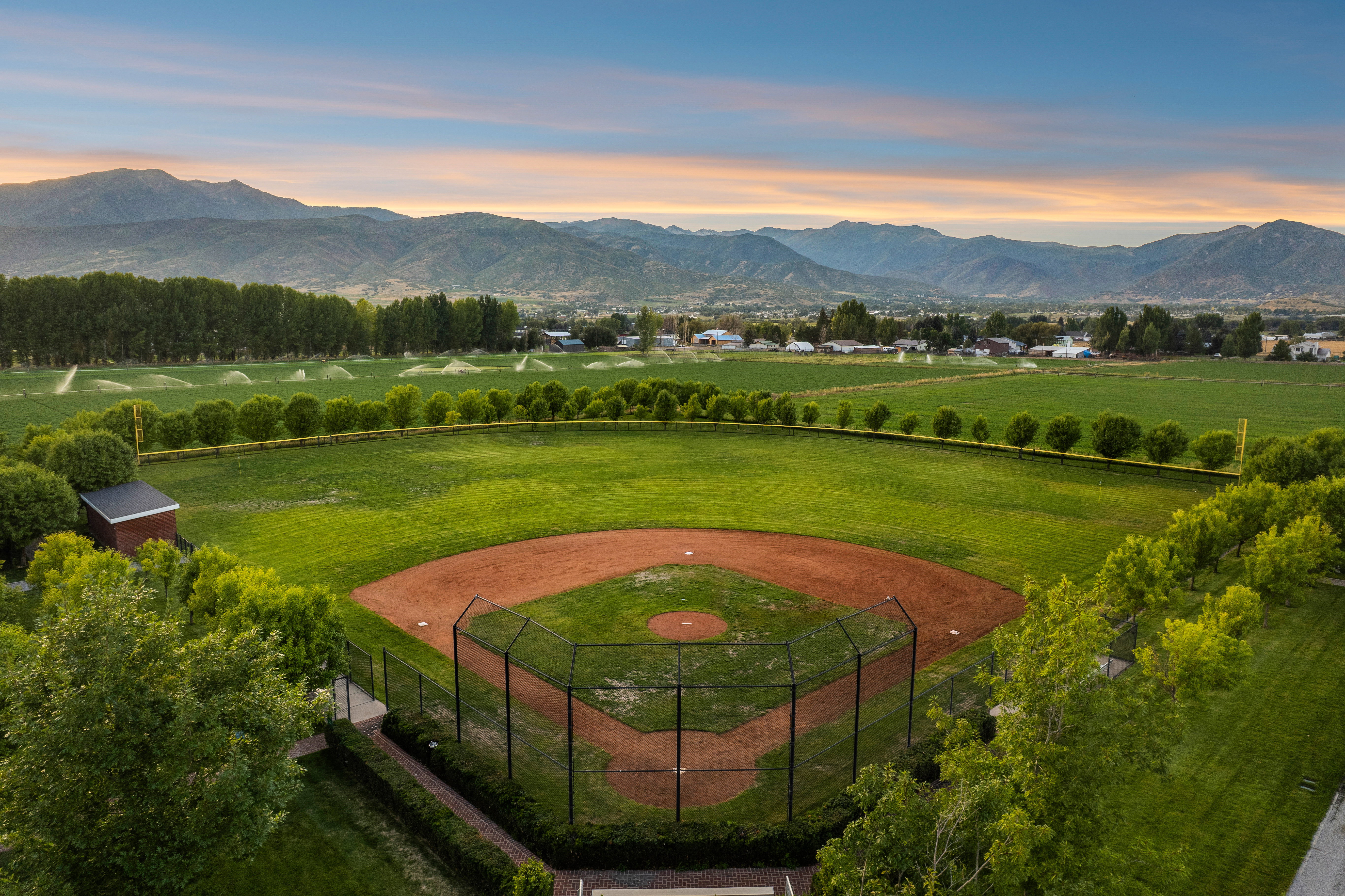 Manicured baseball diamond with a protective backstop, surrounded by lush green fields and mature trees