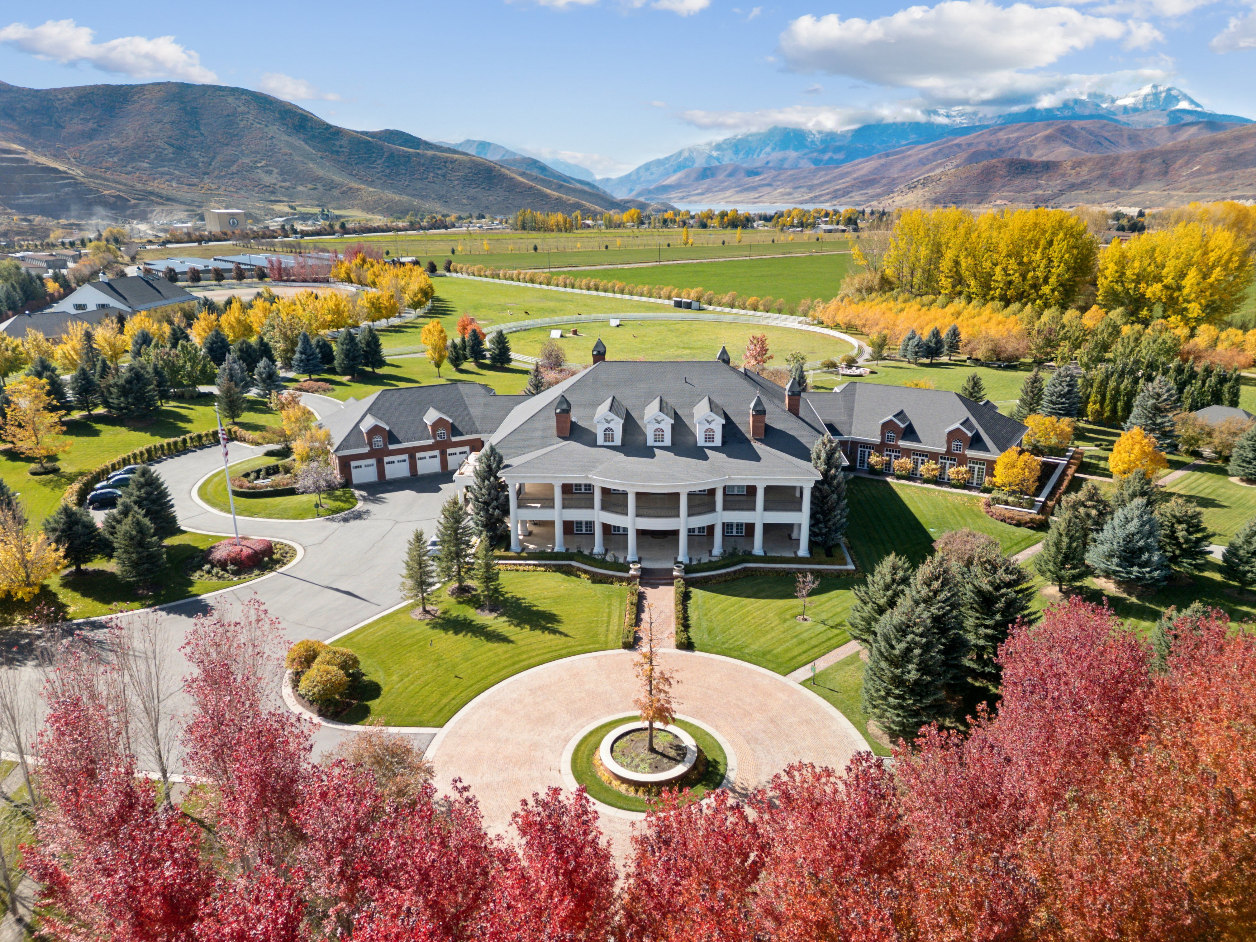 Grand estate featuring a circular drive with a central tree, a covered porch with white columns, multiple dormer windows, and a brick-accented three-car garage
