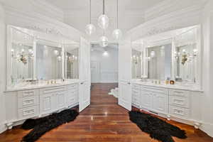 Full bathroom with a decorative wall, dark wood-style flooring, crown molding, two vanities, and a chandelier