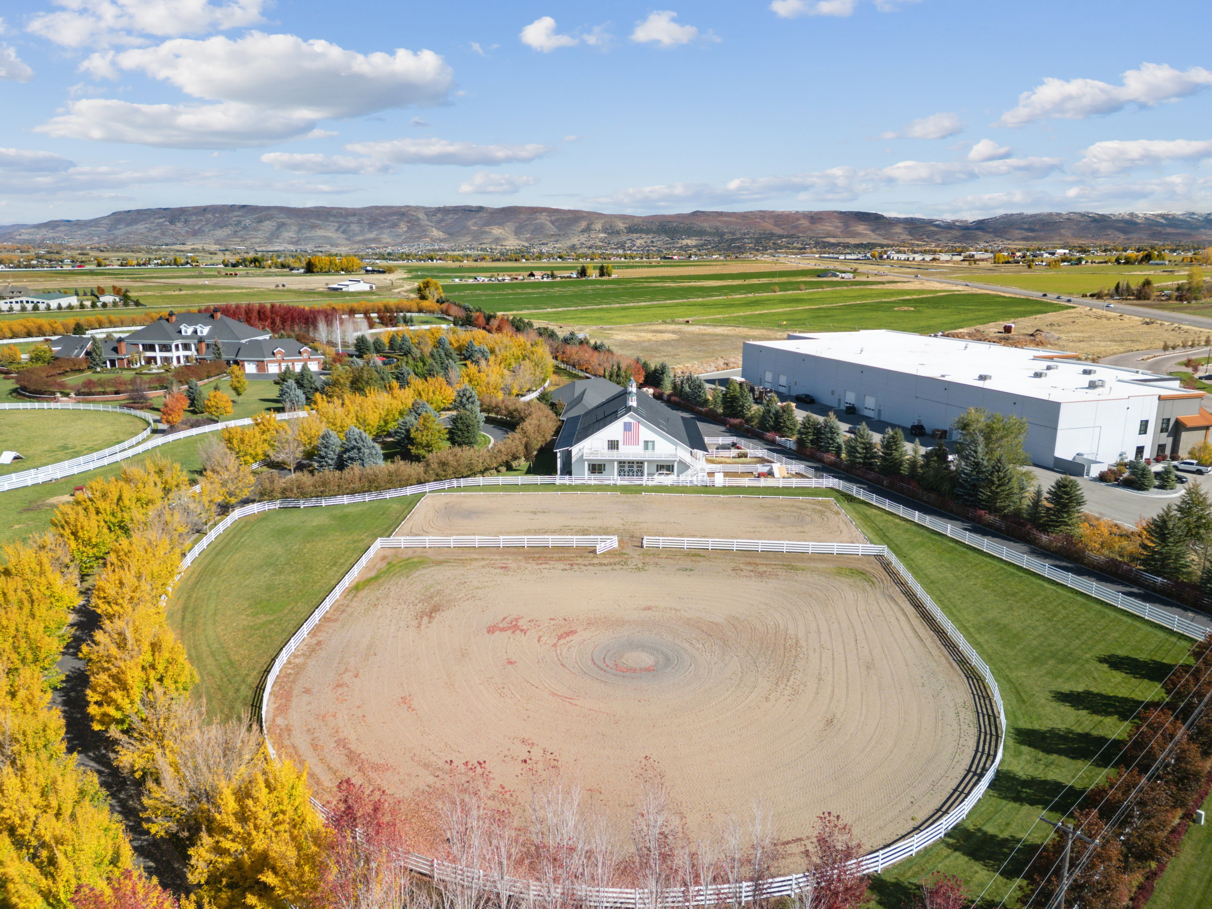 Expansive equestrian arena with white fencing and surrounding green lawns, framed by vibrant autumn foliage