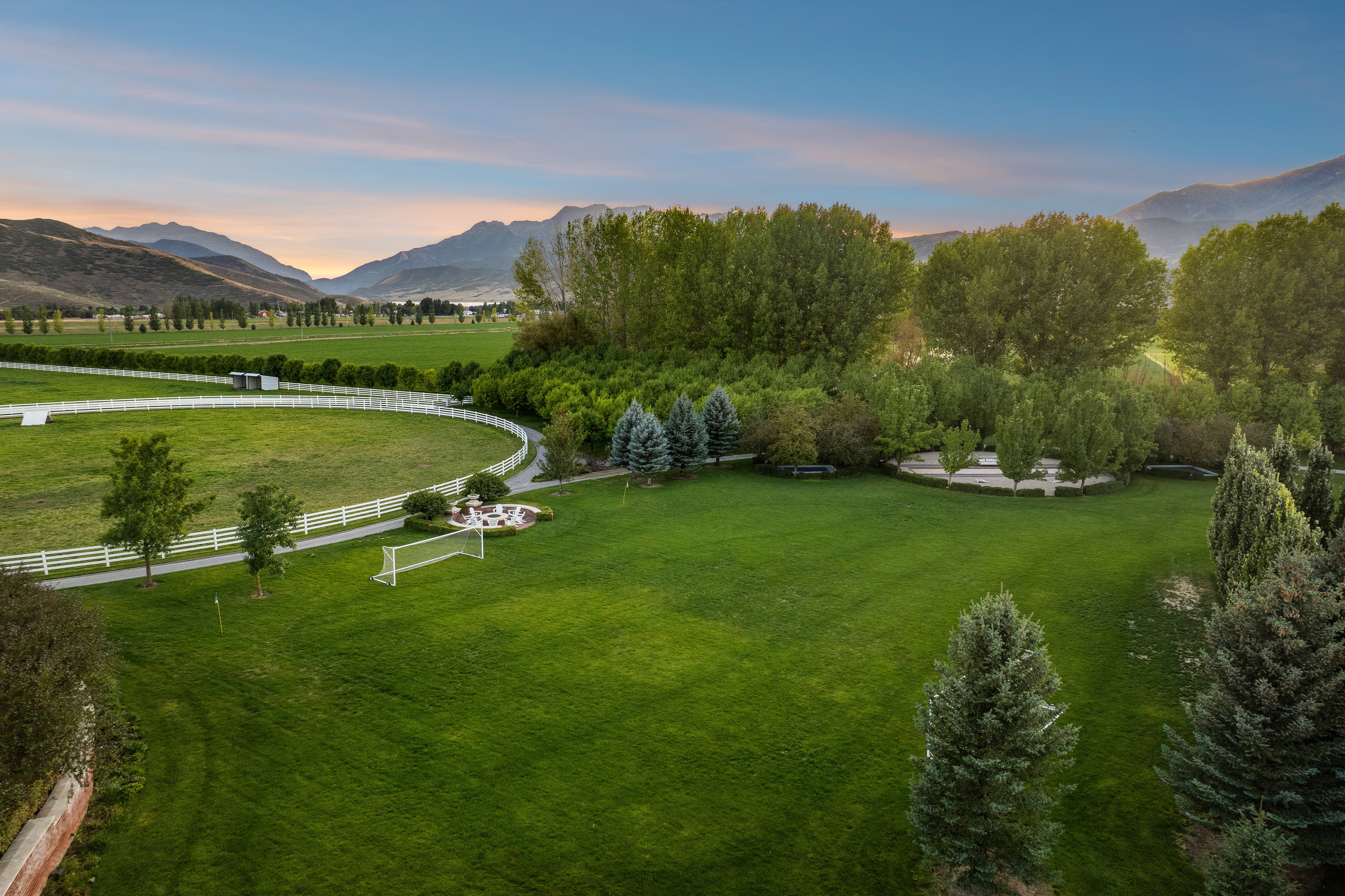 Expansive green lawn surrounded by mature trees, featuring a circular fire pit area with seating, a white rail fence bordering a paved pathway, and distant mountain scenery