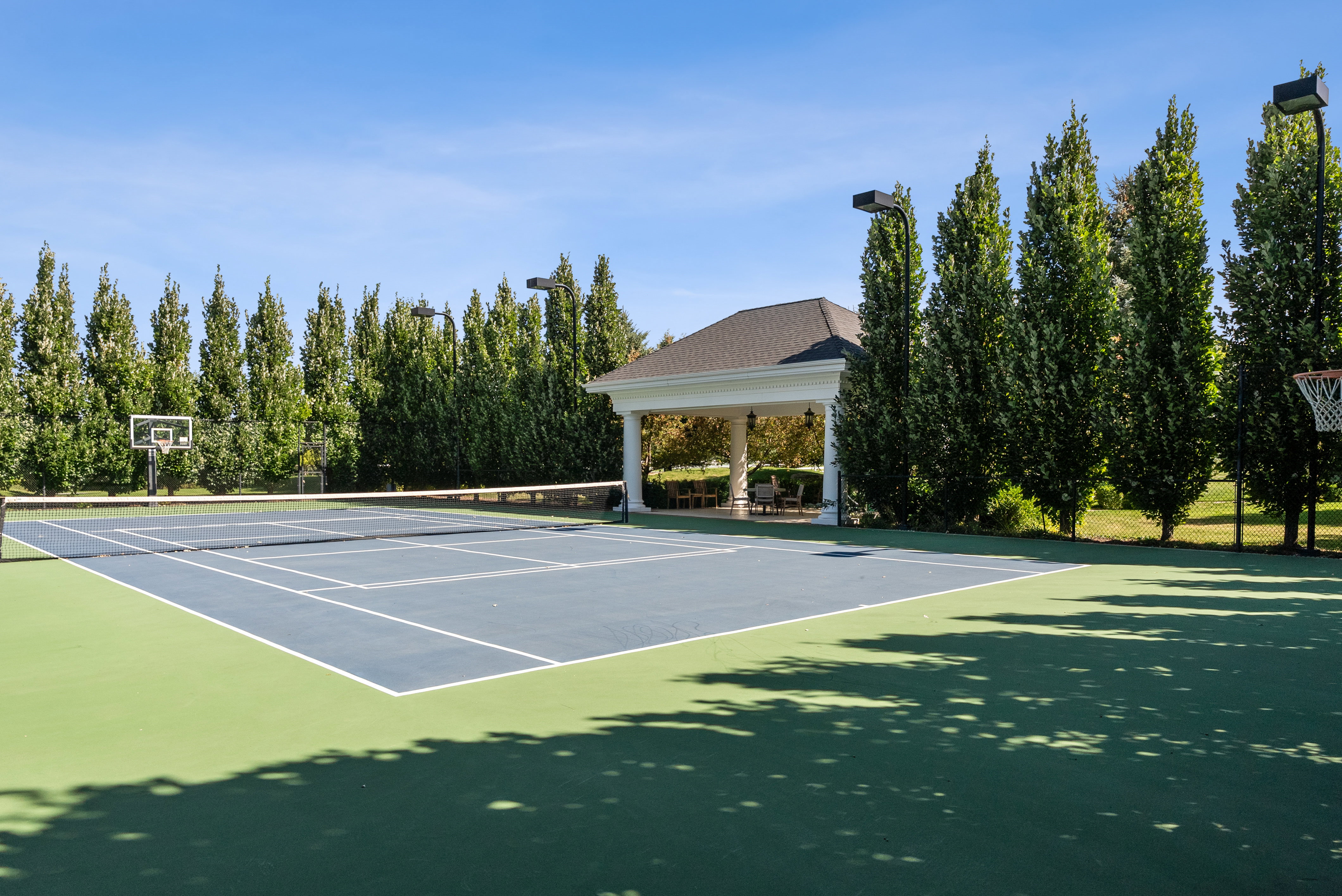 Multi-sport court featuring a tennis net, basketball hoops, and a perimeter of mature trees