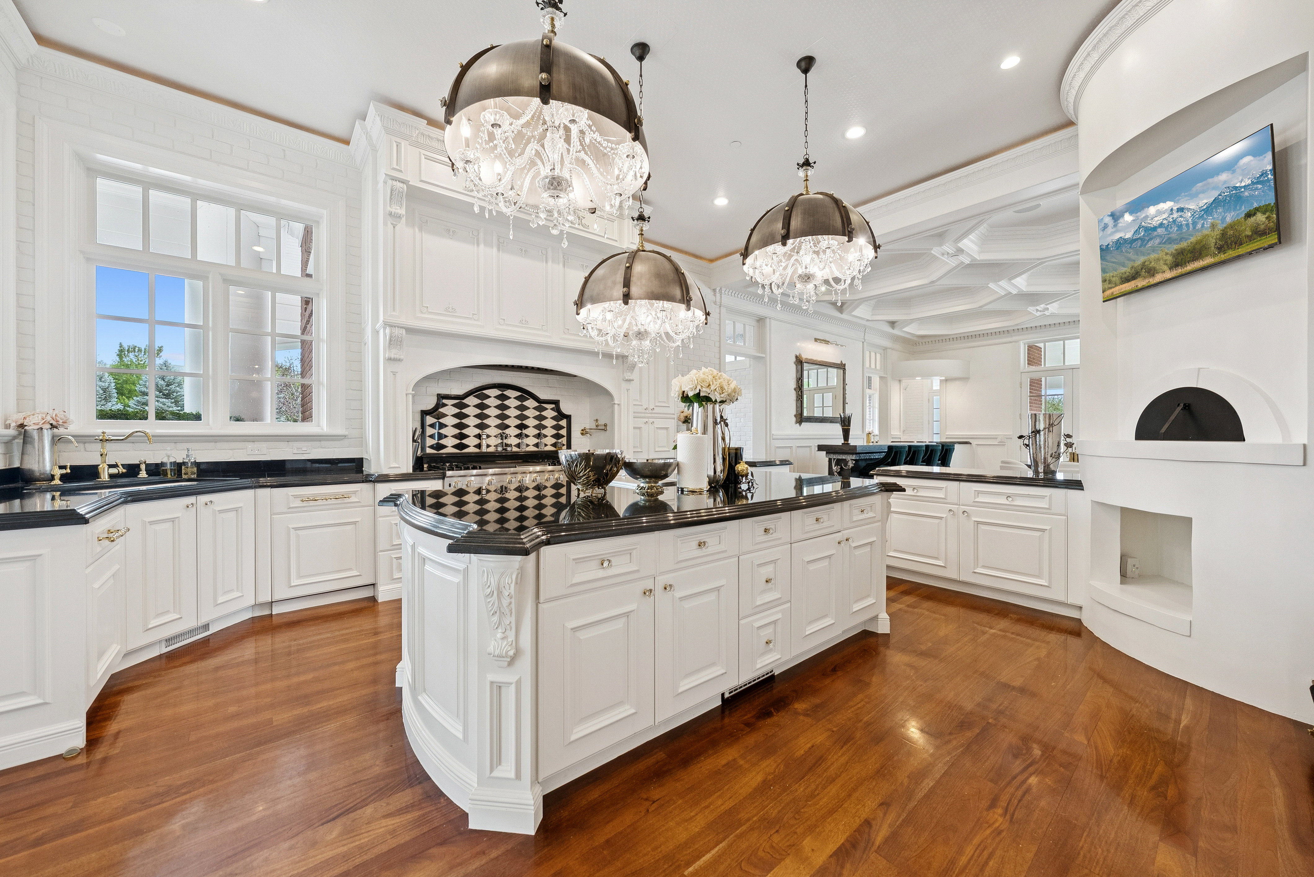 Gourmet kitchen featuring a large central island with black countertops, white custom cabinetry, rich wood-finish flooring, three ornate chandelier light fixtures, and an integrated oven