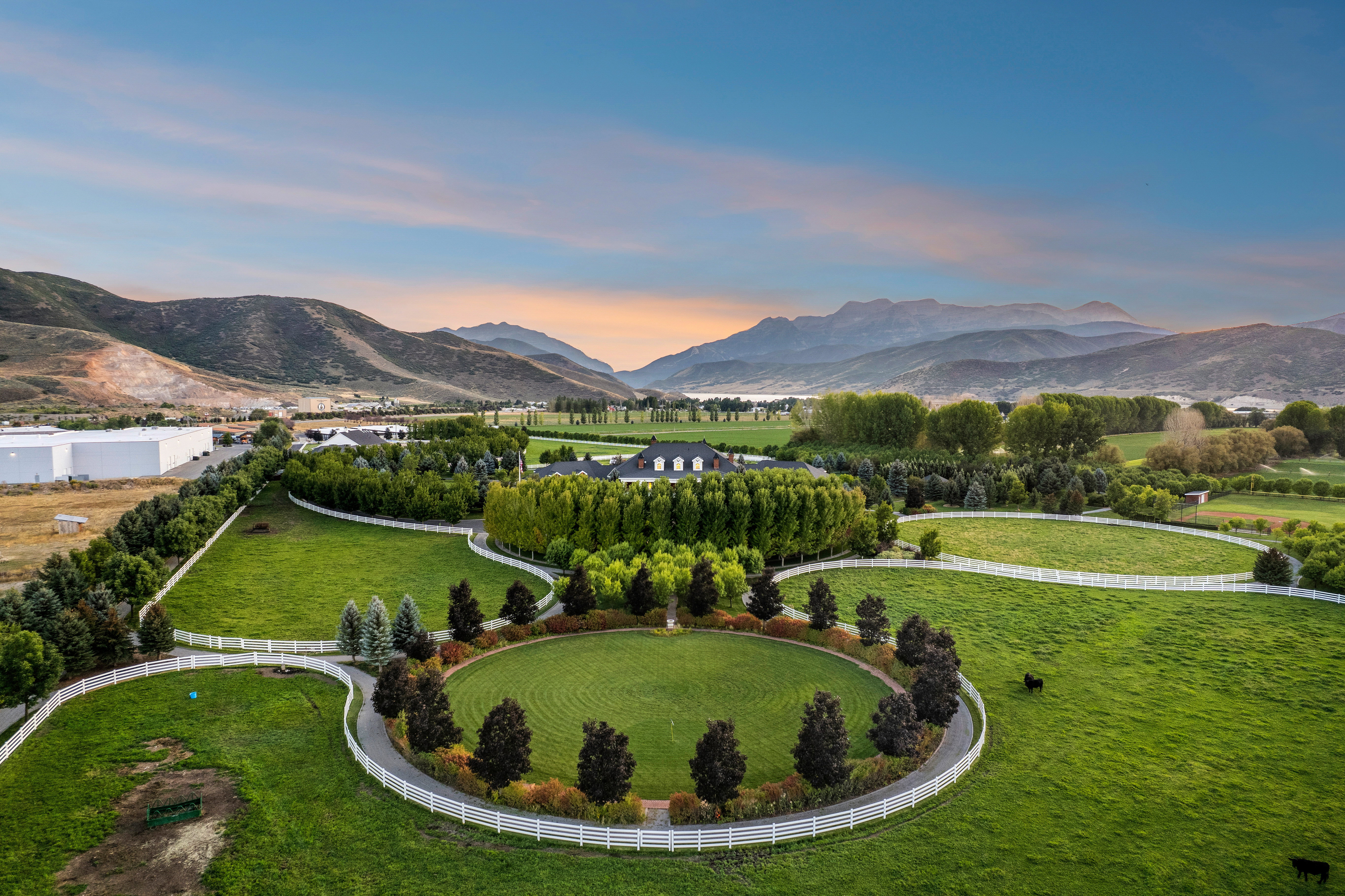 Expansive property featuring manicured green pastures, white fencing, mature trees, a circular driveway, and mountainous scenery