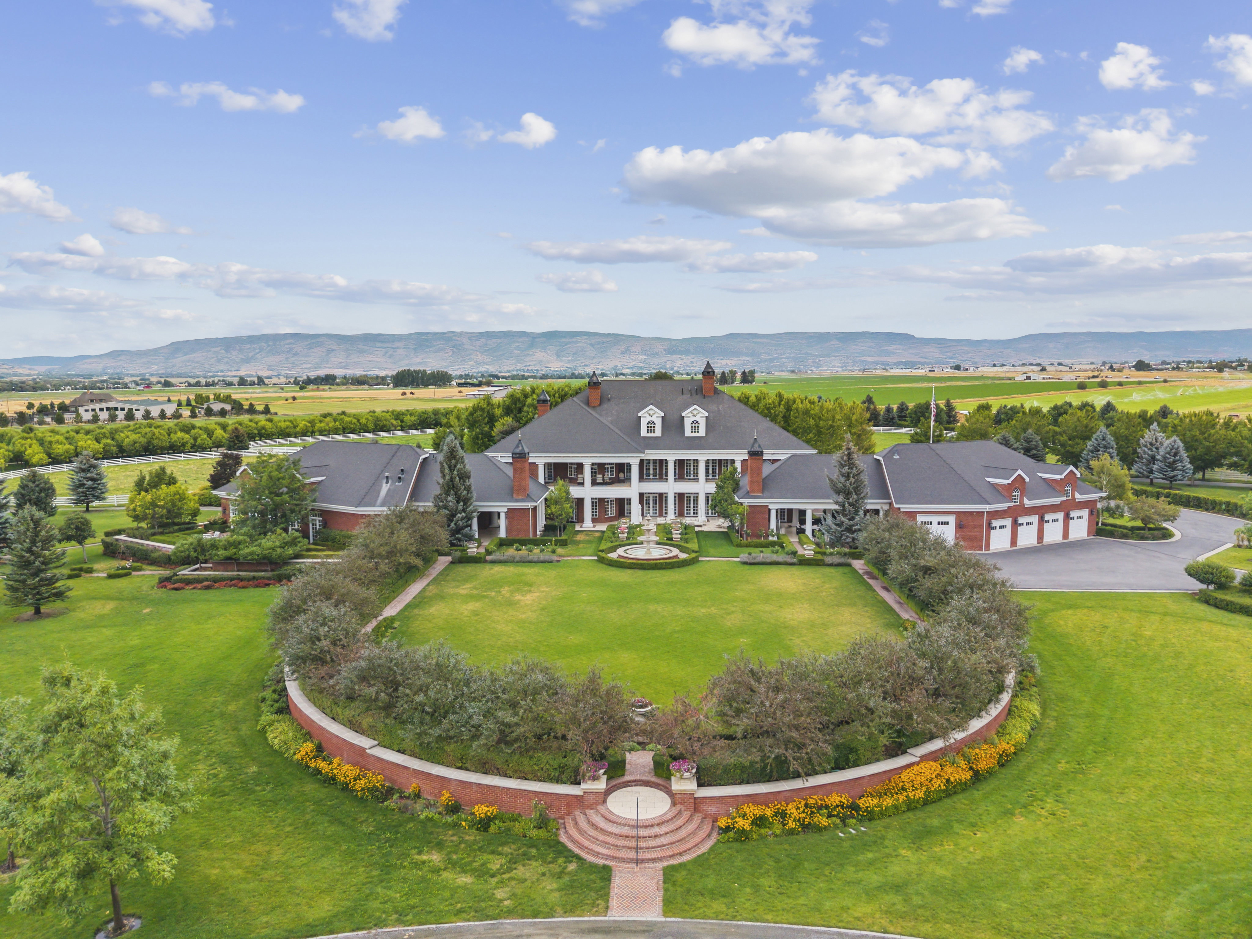 Expansive estate featuring a large main residence with brick exterior and multiple dormer windows, a grand circular driveway, and a separate multi-car garage building