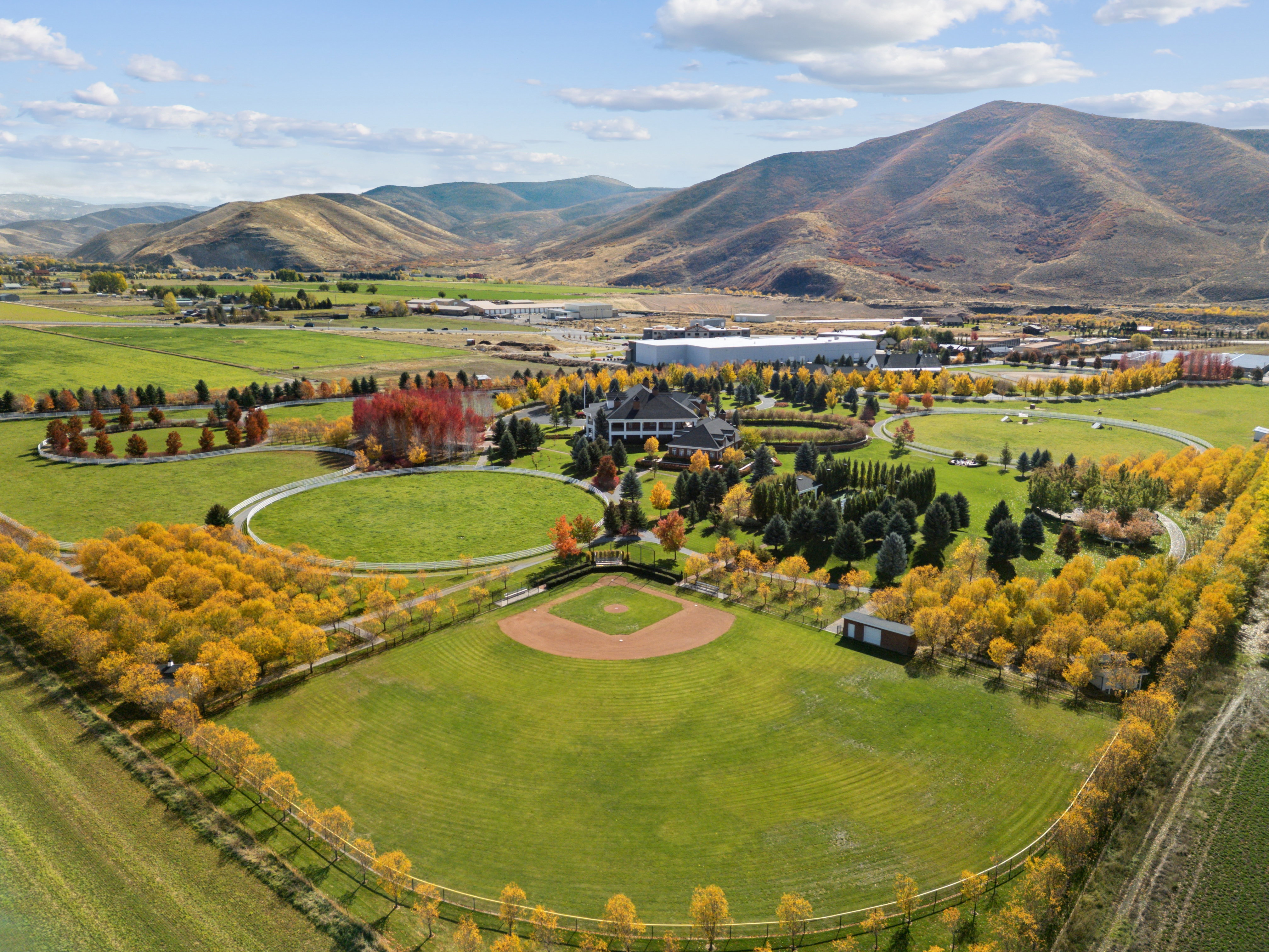 Expansive grounds featuring a baseball diamond, circular lawn area, mature trees, and mountainous terrain in the background