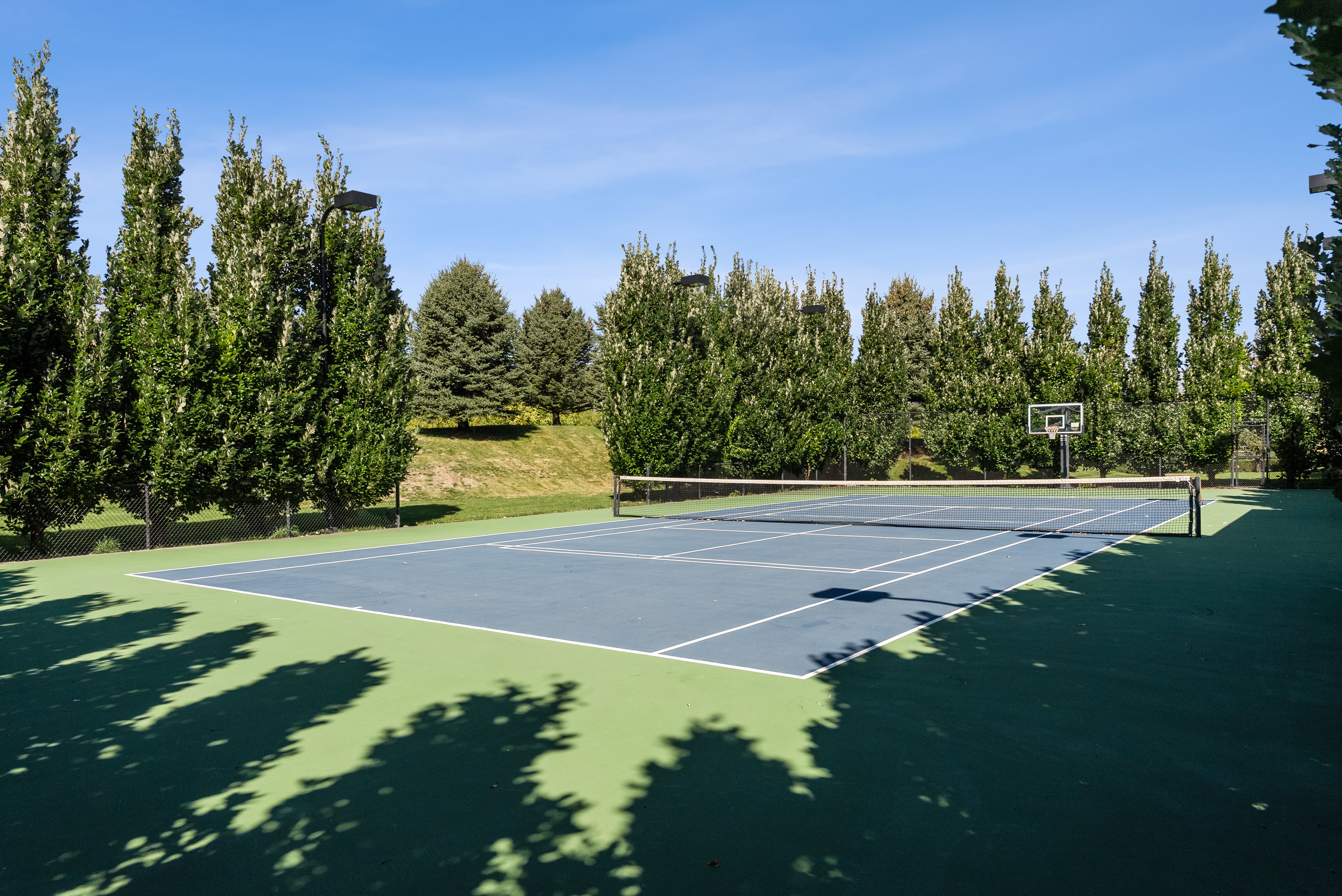 Outdoor tennis court featuring a blue playing surface with white lines, green perimeter, and surrounding mature trees