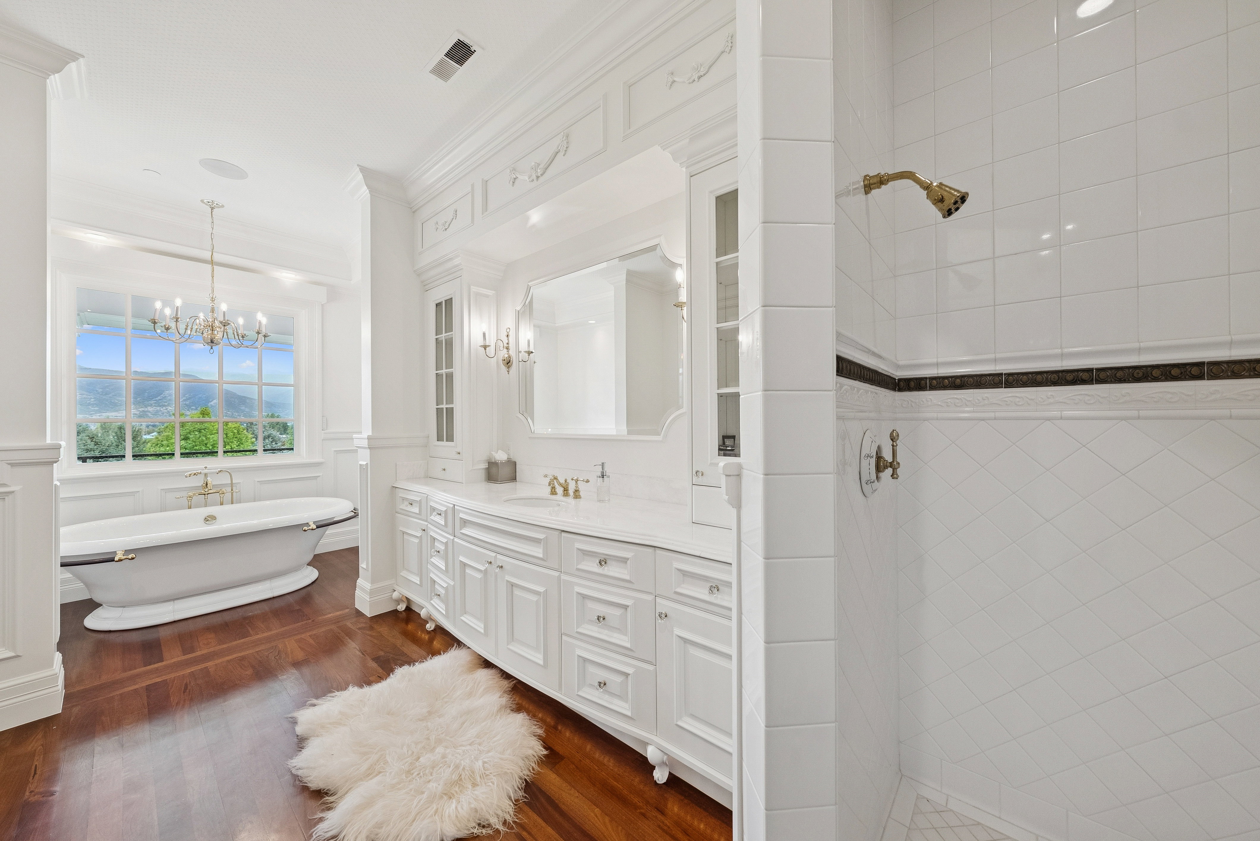 Spacious bathroom featuring a freestanding soaking tub, wood-finish flooring, a double vanity with ample storage, a walk-in shower with tiled walls, and a large window providing expansive scenery
