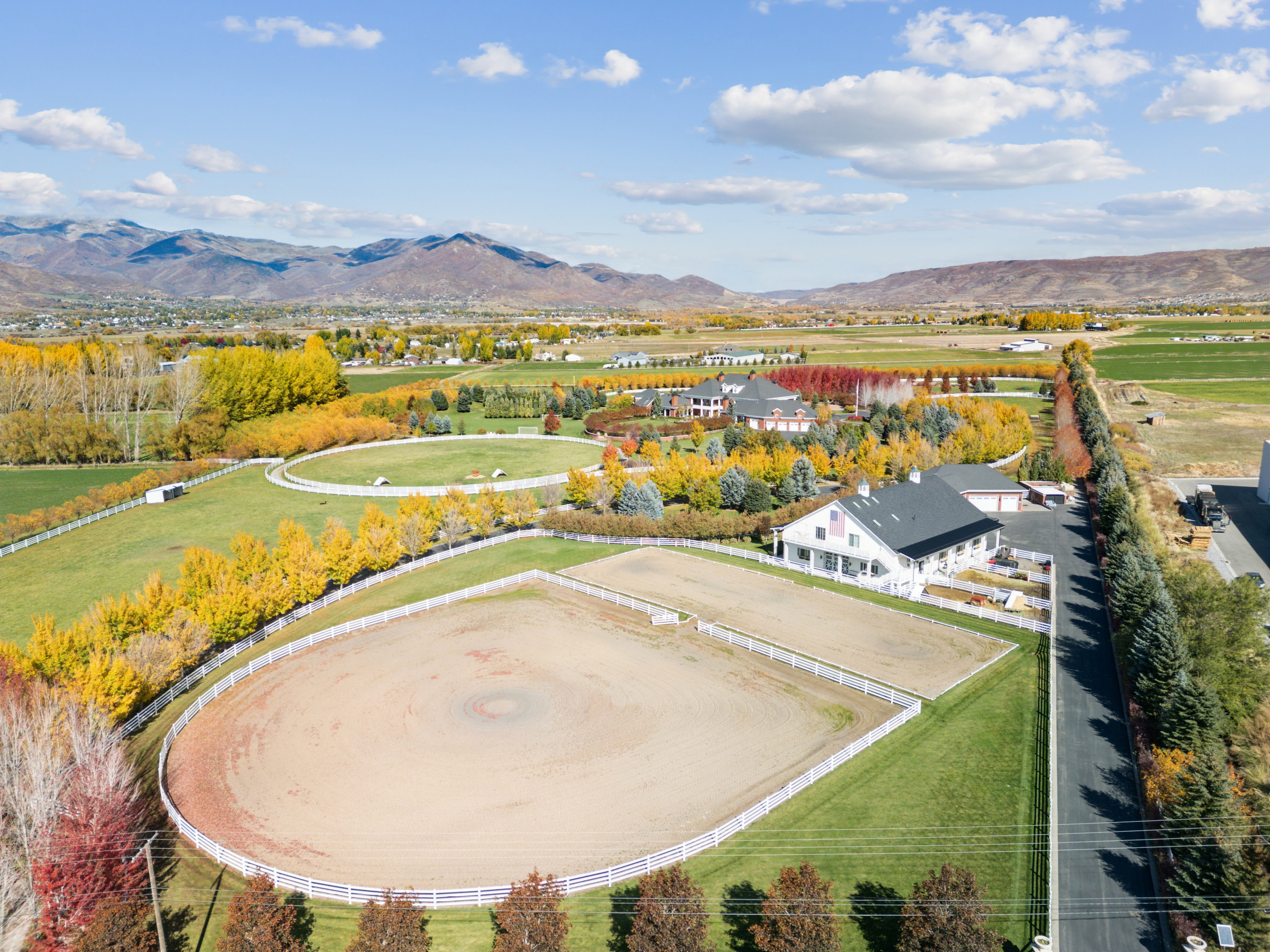 Expansive equestrian property featuring multiple fenced riding arenas, a white farmhouse-style residence with a dark roof, a detached garage, and mature landscaping with vibrant autumn foliage