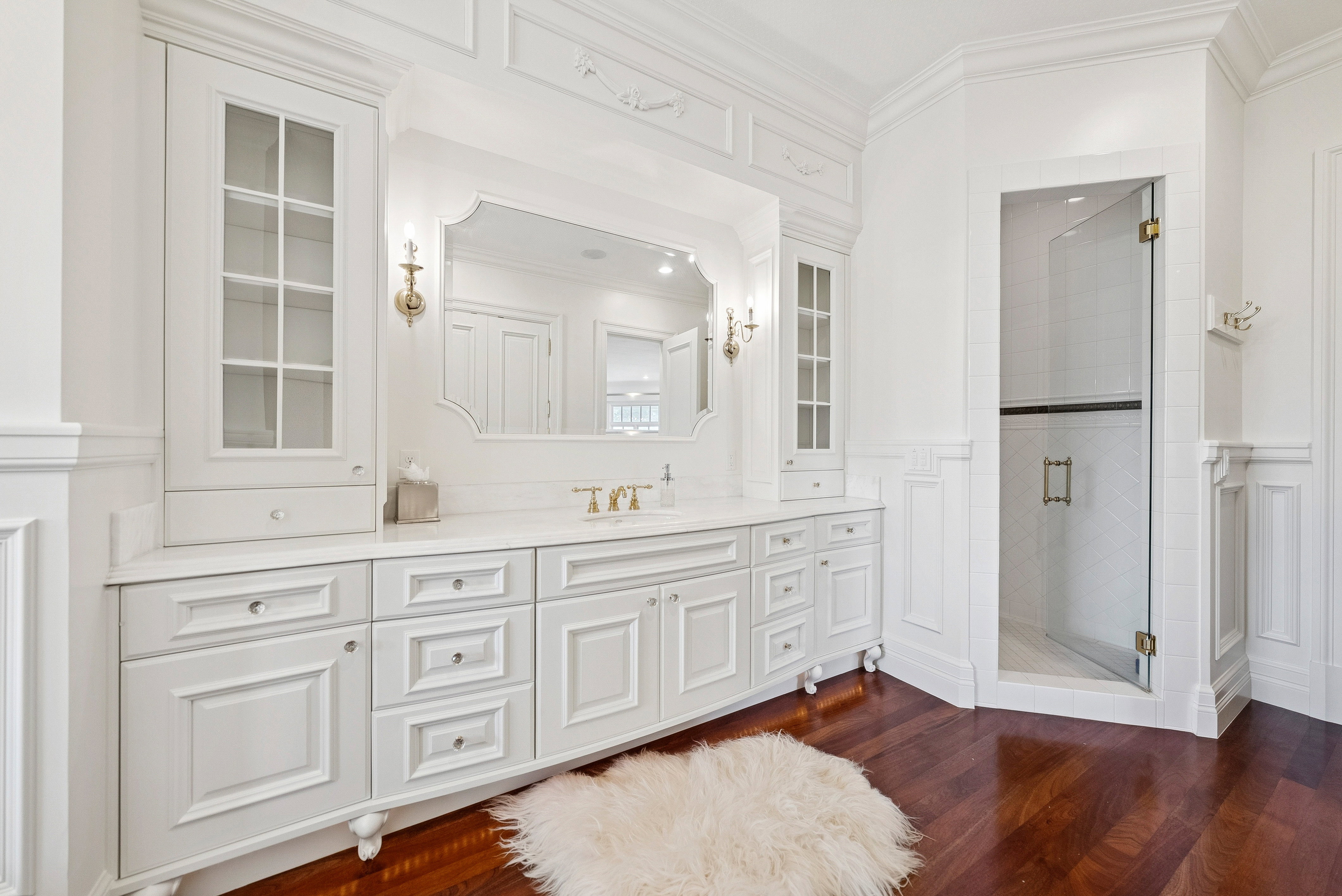 White custom cabinetry with glass-front display cabinets, a recessed vanity mirror, and dual wall sconces