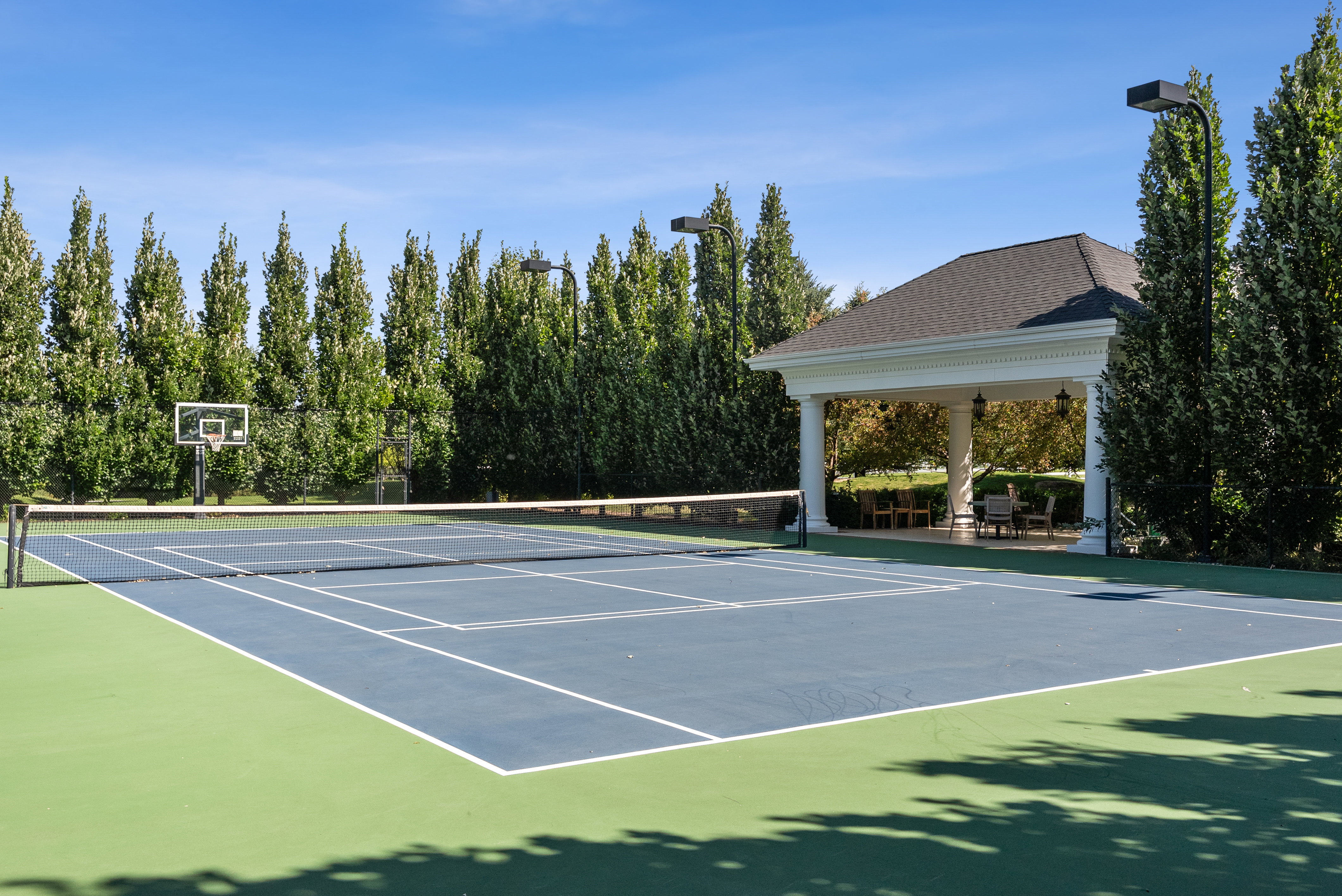 Full-size tennis court with basketball hoop, surrounded by mature trees