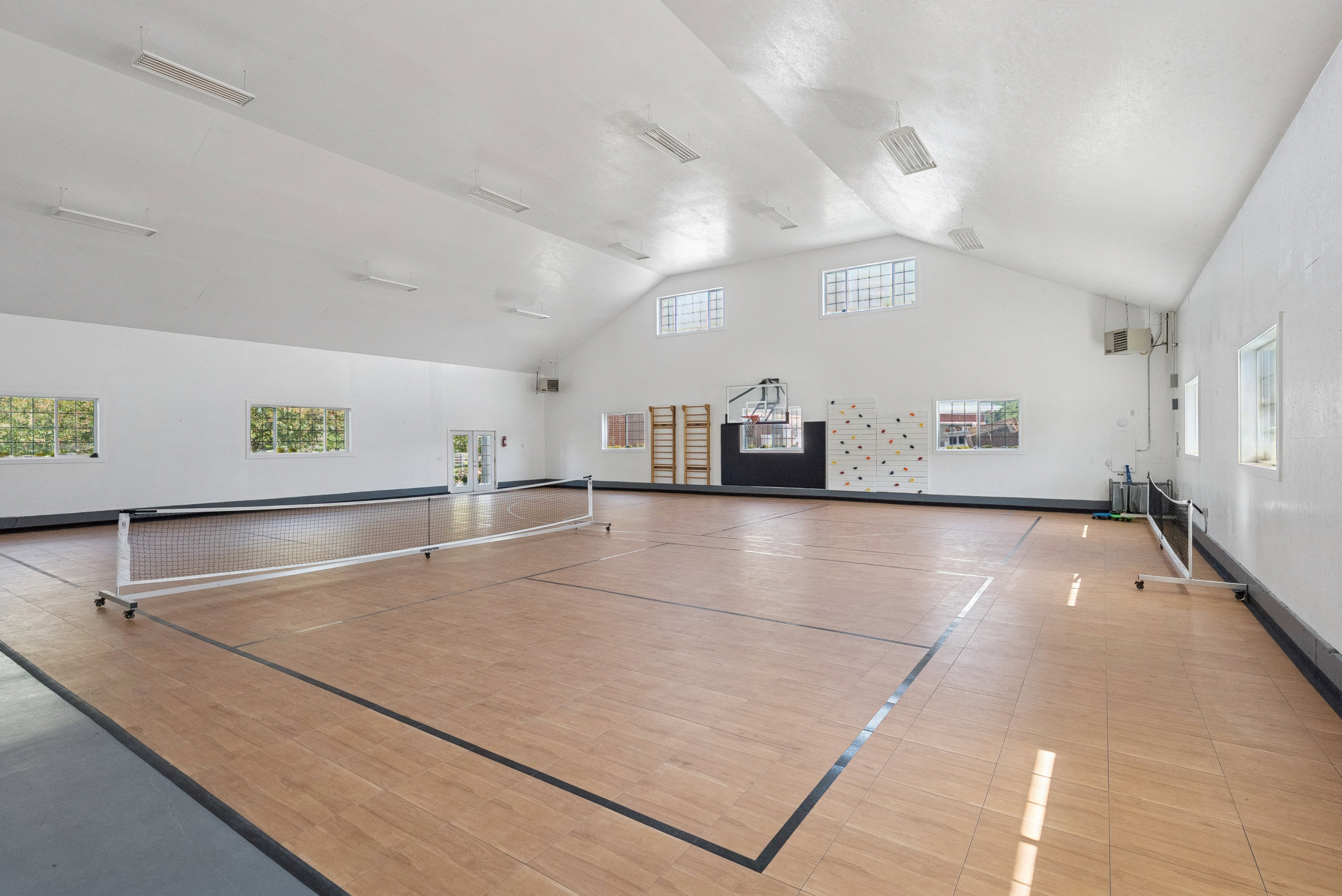 Spacious interior athletic court featuring wood-finish flooring with court lines, a full-height ceiling, multiple windows, and recessed lighting