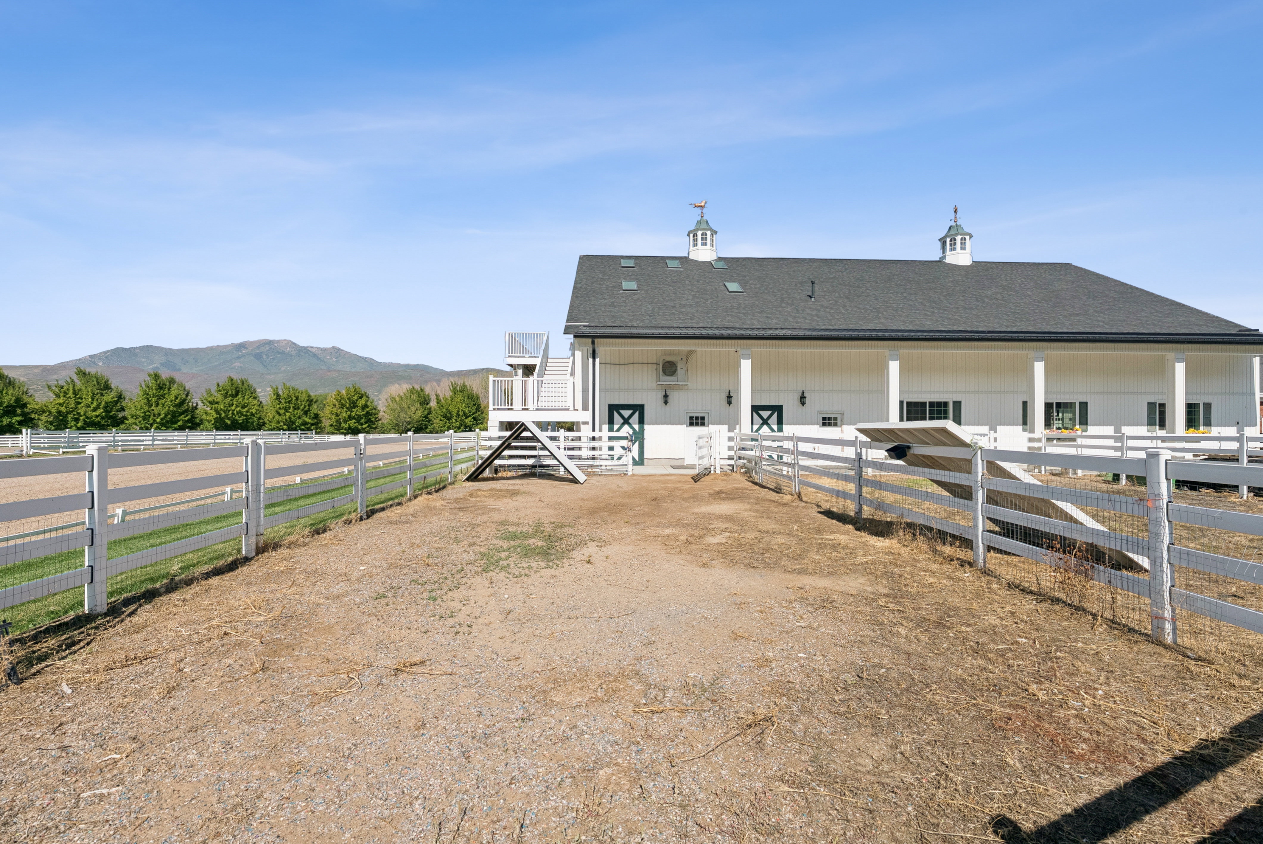White barn structure featuring a dark shingle roof, two cupolas with weather vanes, and a surrounding white fence