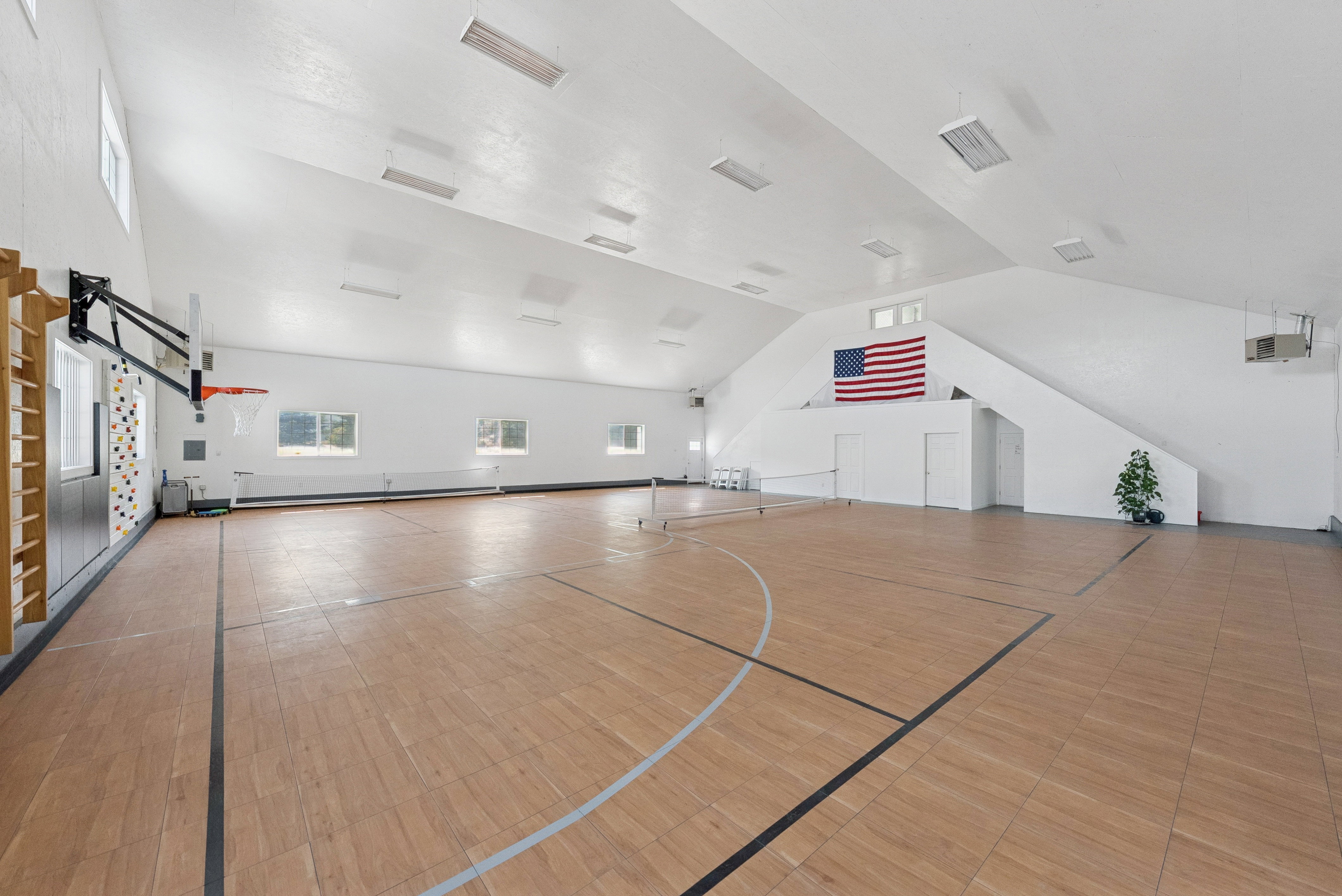 Spacious indoor court featuring wood-finish flooring with marked lines, a basketball hoop, and a net for other sports