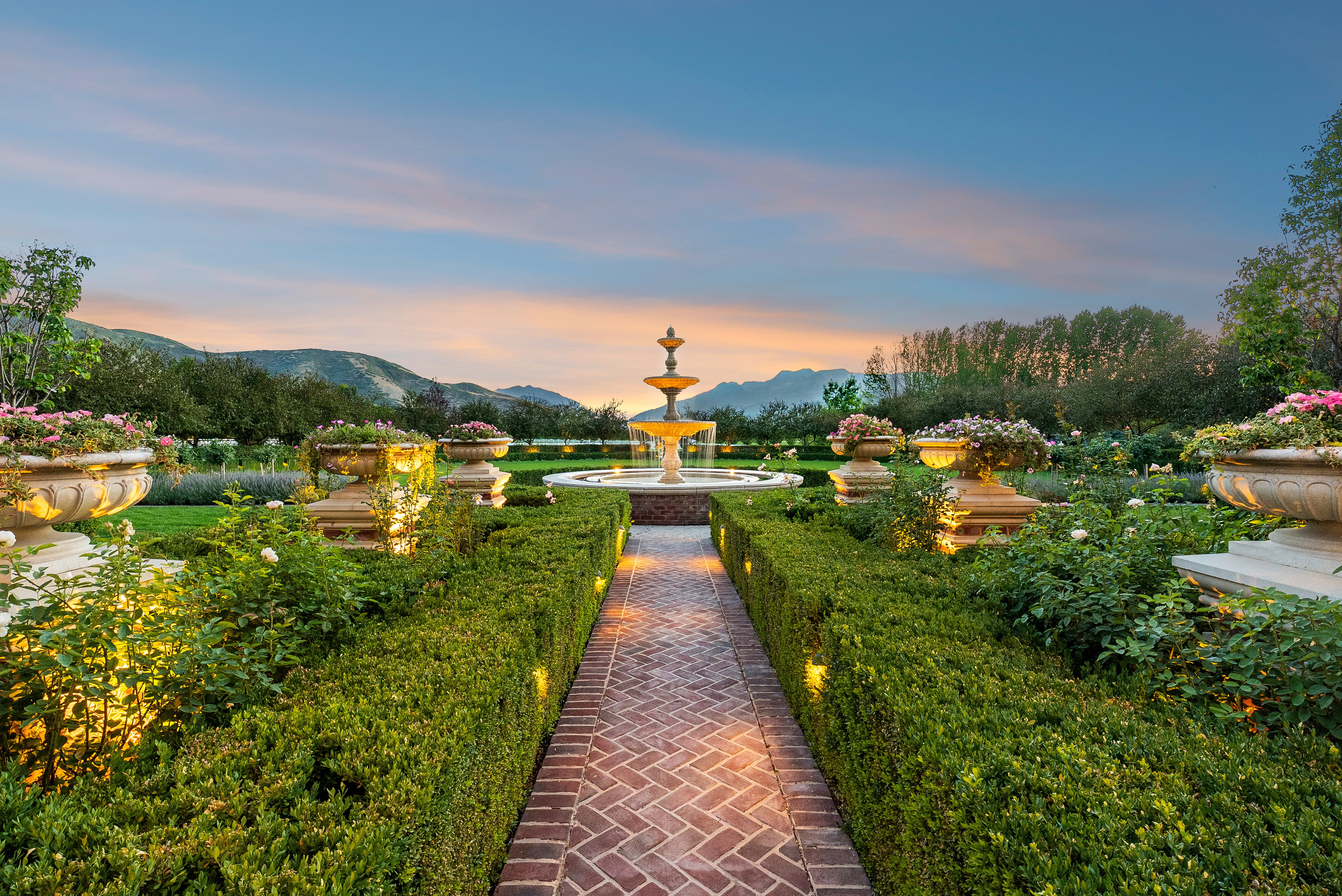 Expansive garden featuring a tiered fountain, intricate brick pathway, manicured hedges, and decorative urns with plantings