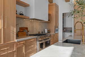 Kitchen with tasteful backsplash, range with two ovens, wall chimney exhaust hood, brown cabinets, and light stone counters