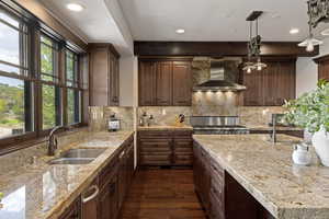 Kitchen with dark wood-type flooring, wall chimney range hood, light stone countertops, backsplash, and dark brown cabinetry